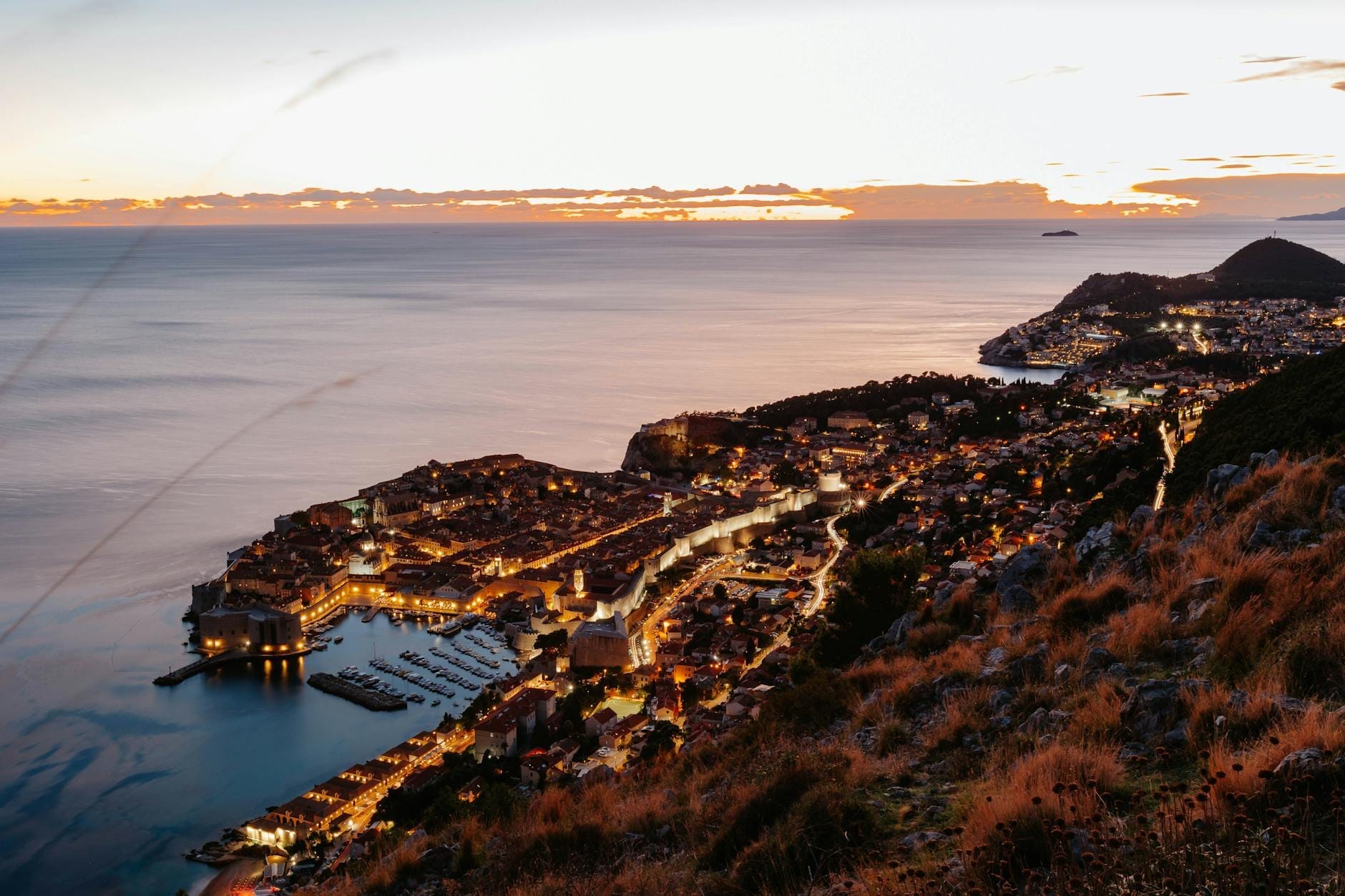 Vista aérea impresionante de Dubrovnik al atardecer, con las murallas iluminadas de la ciudad antigua y el mar Adriático de fondo.