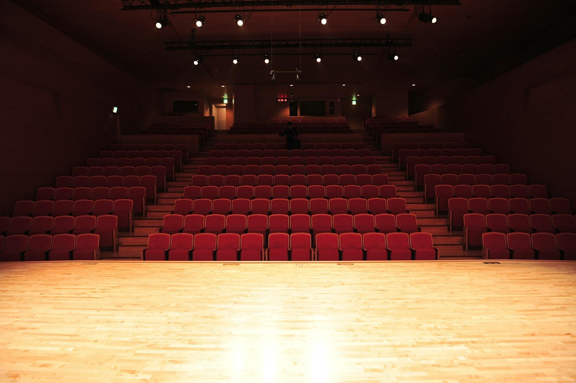 Teatro vacío con butacas rojas y escenario iluminado por focos, visto desde el escenario hacia el patio de butacas.