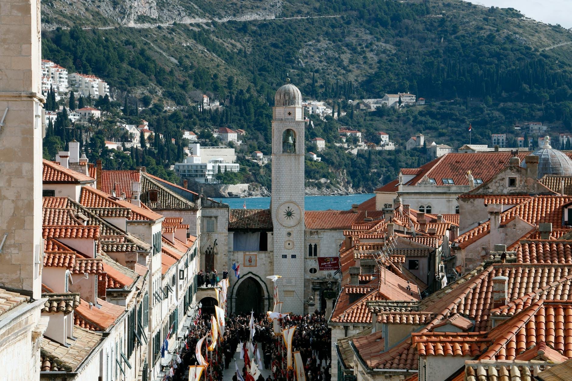 Vista panorámica de la Ciudad Vieja de Dubrovnik con multitudes entre edificios históricos de piedra, la torre del reloj, las murallas de la ciudad y el mar de fondo en un día soleado.