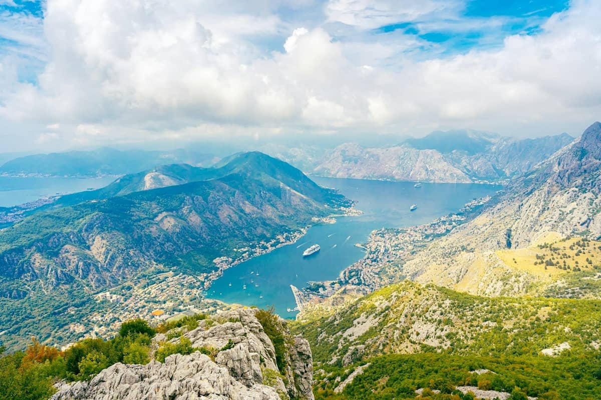 Panoramic view of the Bay of Kotor with winding coastal road, mountains, and cruise ships in the bay below.