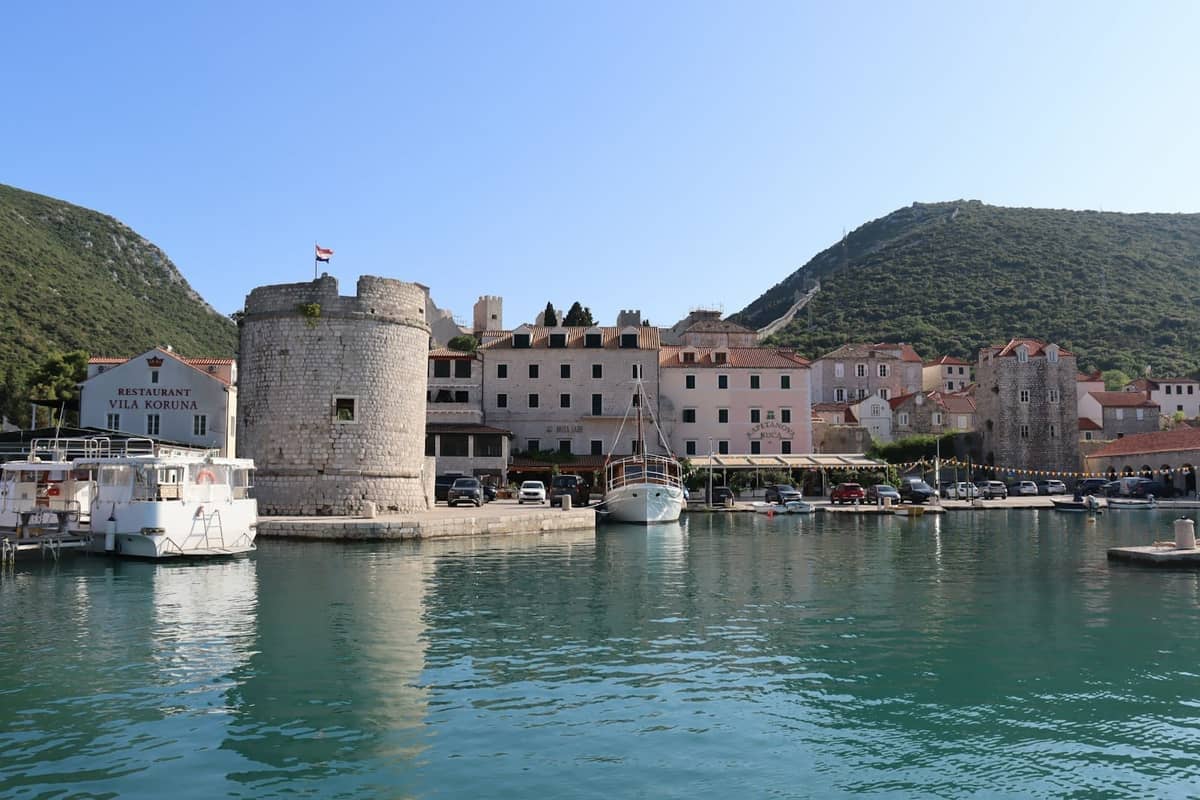 Scenic view of Kotor old town's waterfront with medieval tower, historic buildings, boats docked in the harbor, and green hills in the background.