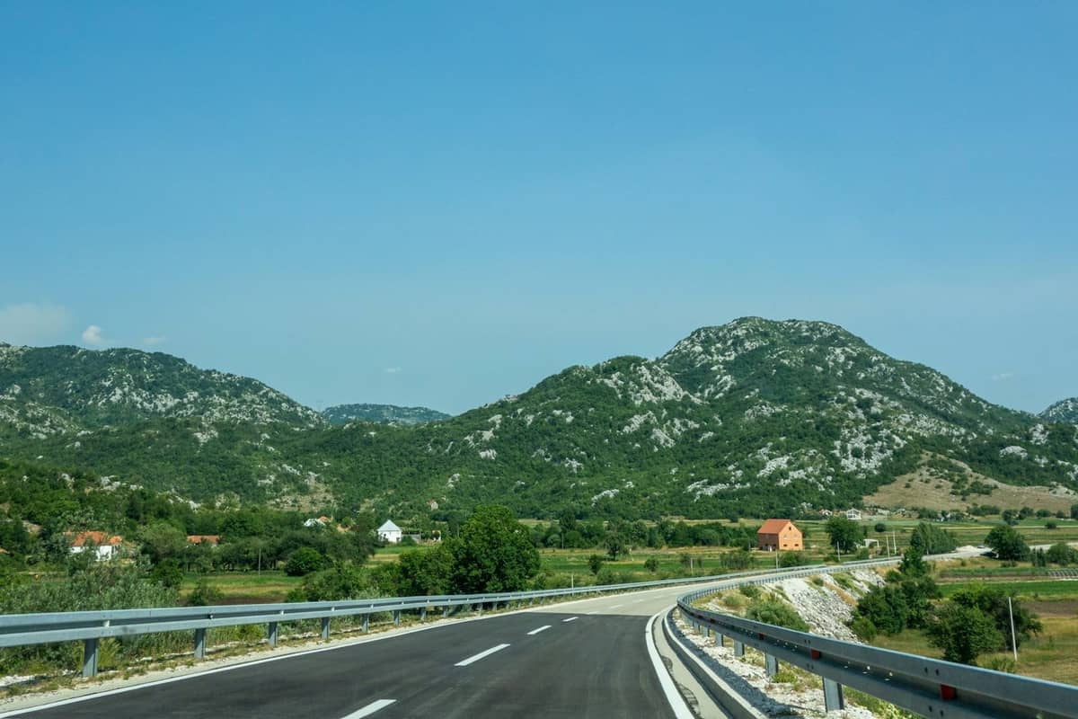 A smooth asphalt road with guardrails curves toward the green hills and mountains, evoking the drive to a border crossing.