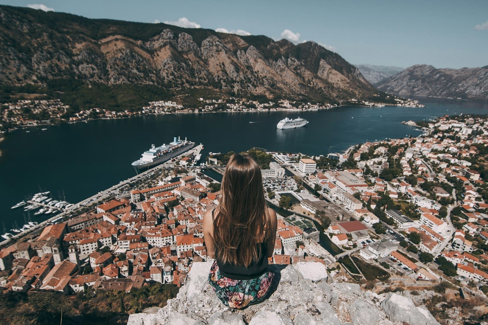 Uma mulher sentada sobre rochas com vista para a Baía de Kotor, com telhados de terracota, navios de cruzeiro e montanhas dramáticas sob um céu brilhante ao meio-dia.