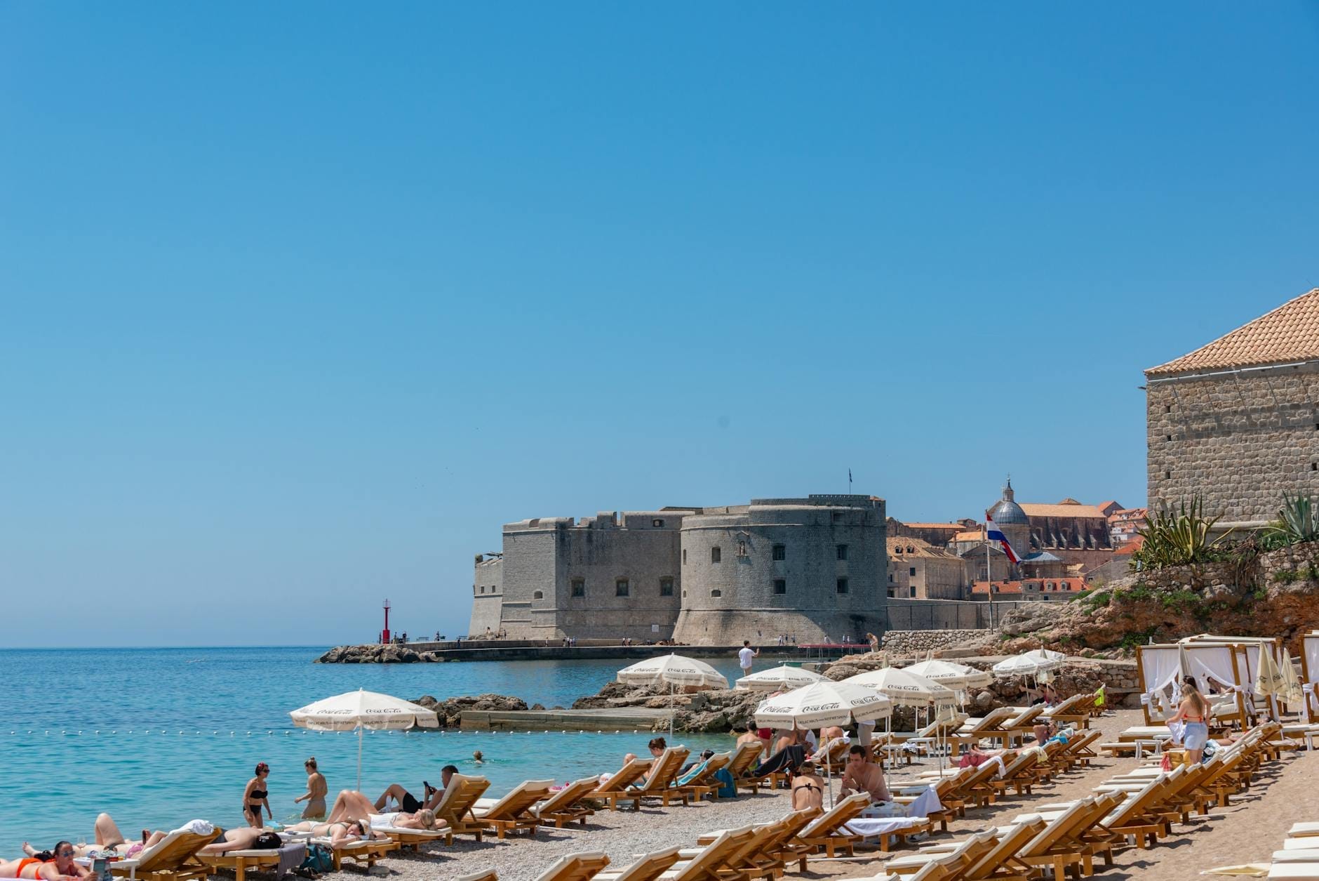 Vue des remparts de la vieille ville de Dubrovnik avec des baigneurs sur une plage de sable peu fréquentée avec des parasols et une mer bleue cristalline sous un ciel ensoleillé.