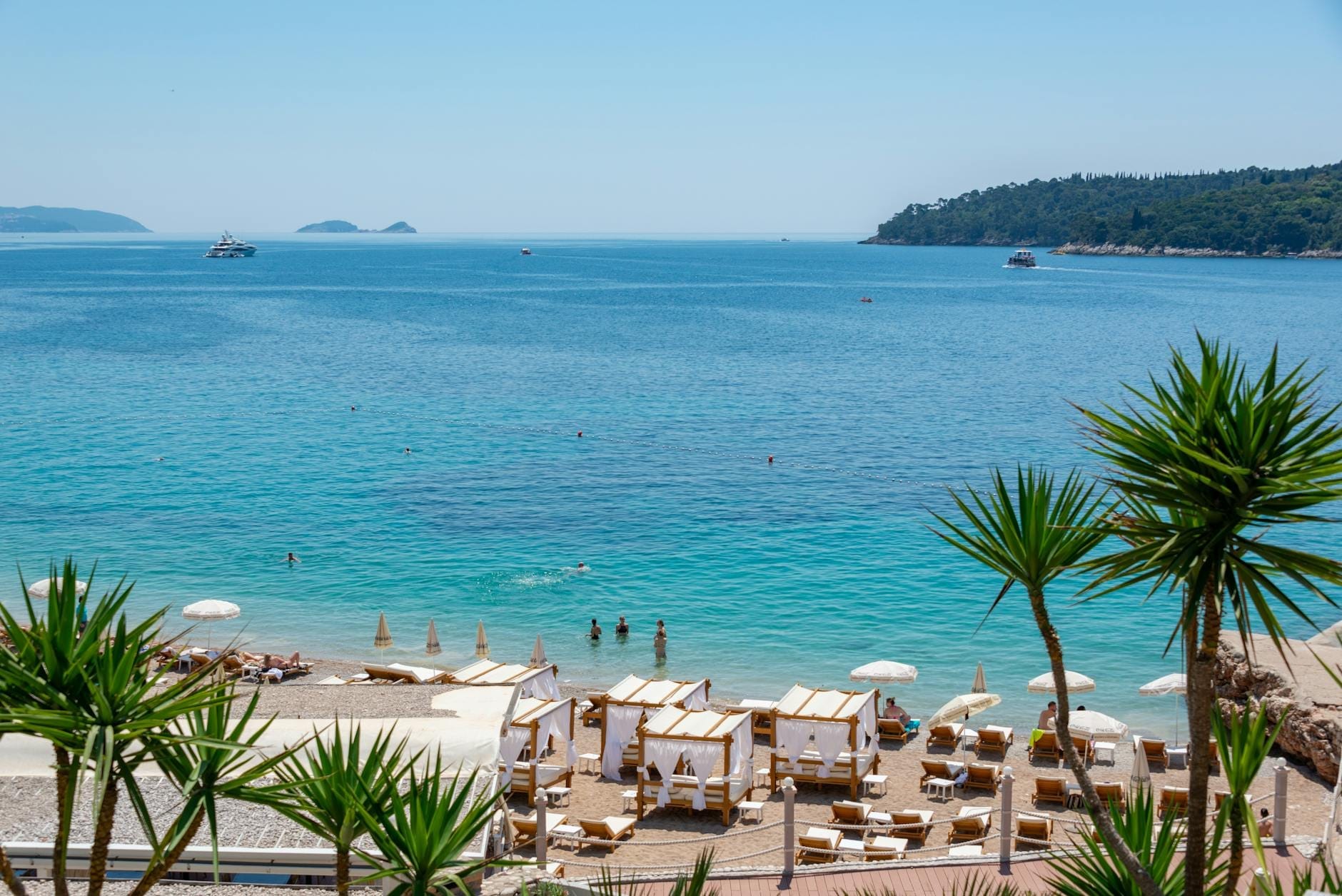 Plage ensoleillée de Dubrovnik avec des cabanes et des personnes nageant dans la mer Adriatique bleue et cristalline, avec des bateaux au loin et une luxuriante île verdoyante en toile de fond.