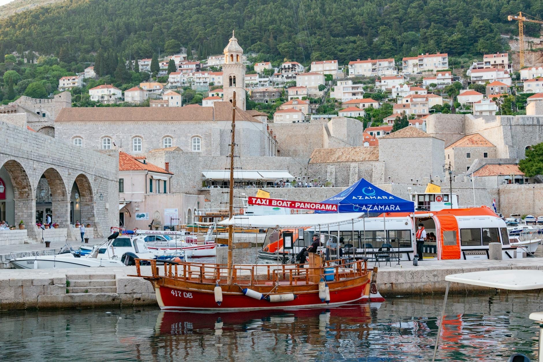 Tekneler, görünür bir su taksisi tabelası ve arka planda şehir surlarıyla Dubrovnik limanı, yerel ulaşım seçeneklerini gösteriyor.