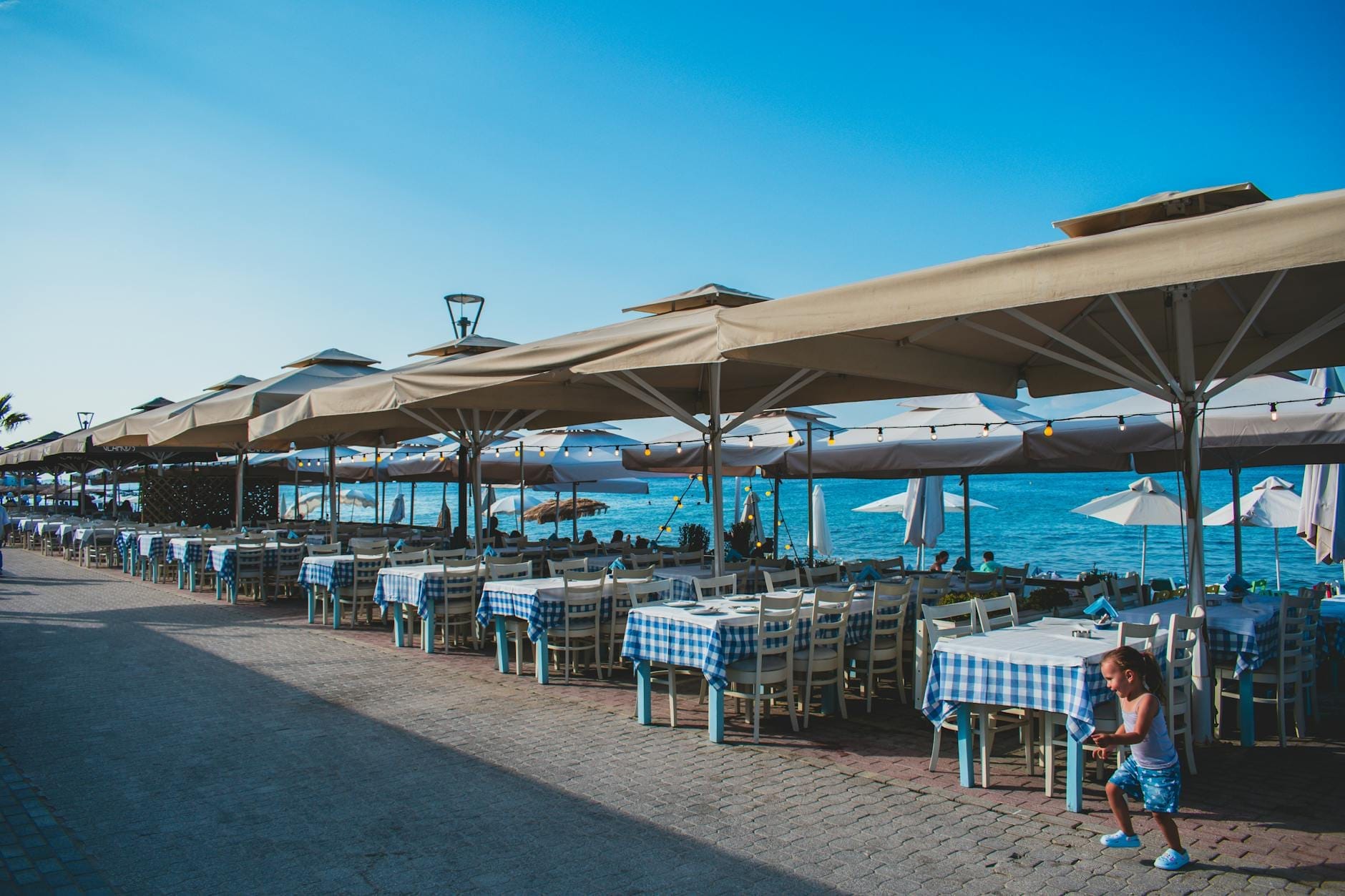 Dubrovnik'te mavi-beyaz masa örtüleri, güneş şemsiyeleri ve açık mavi gökyüzü altında masaların yanında bir çocuk olan deniz kenarı açık hava restoranı.