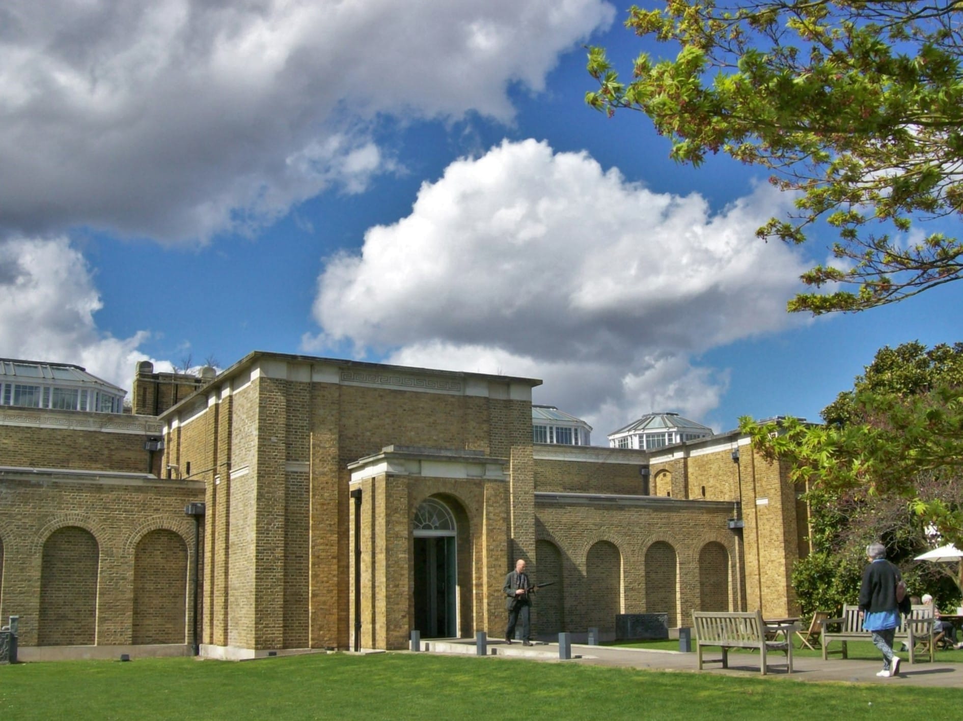 La maestosa facciata in mattoni gialli della Dulwich Picture Gallery sotto un cielo nuvoloso e drammatico, con panchine e visitatori che passeggiano in primo piano.