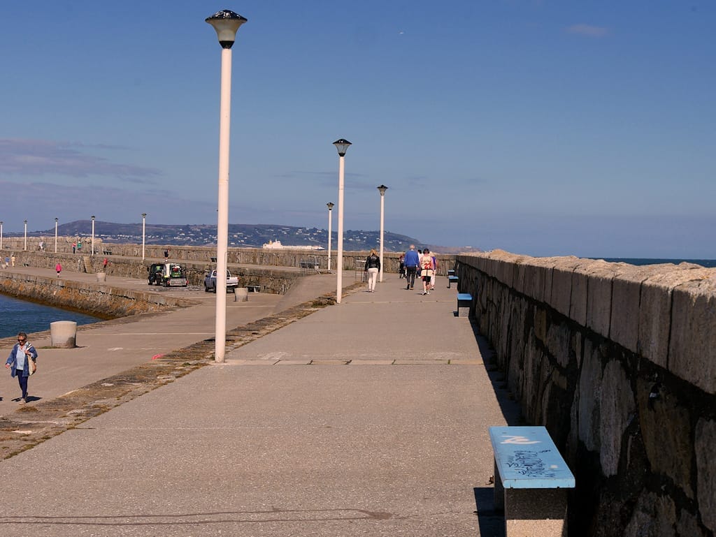 Vista ampia del molo est di Dún Laoghaire con passeggiata in pietra, lampioni, panchine e persone che passeggiano lungo la costa sotto un cielo azzurro limpido.