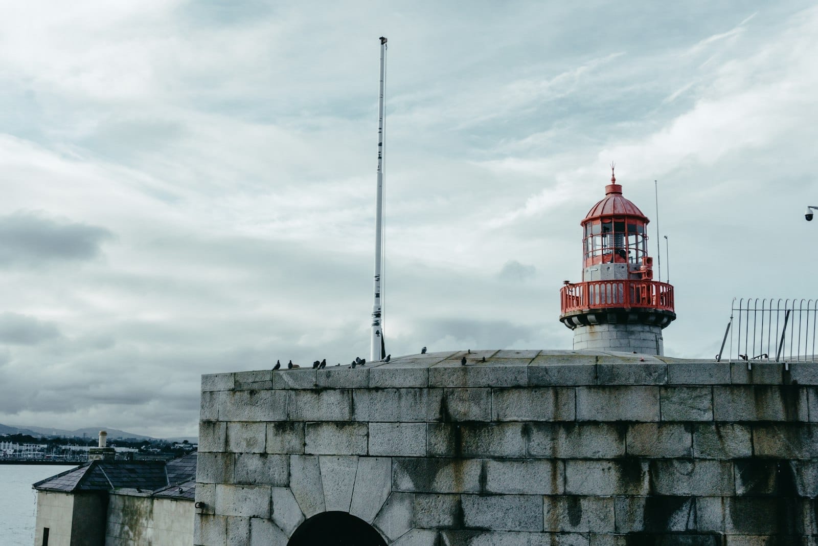 ท่าเรือหินและประภาคารสีแดงประวัติศาสตร์ที่ท่าเรือ Dún Laoghaire ใต้ท้องฟ้าครึ้ม พร้อมนกเกาะอยู่บนกำแพงที่ผุพัง
