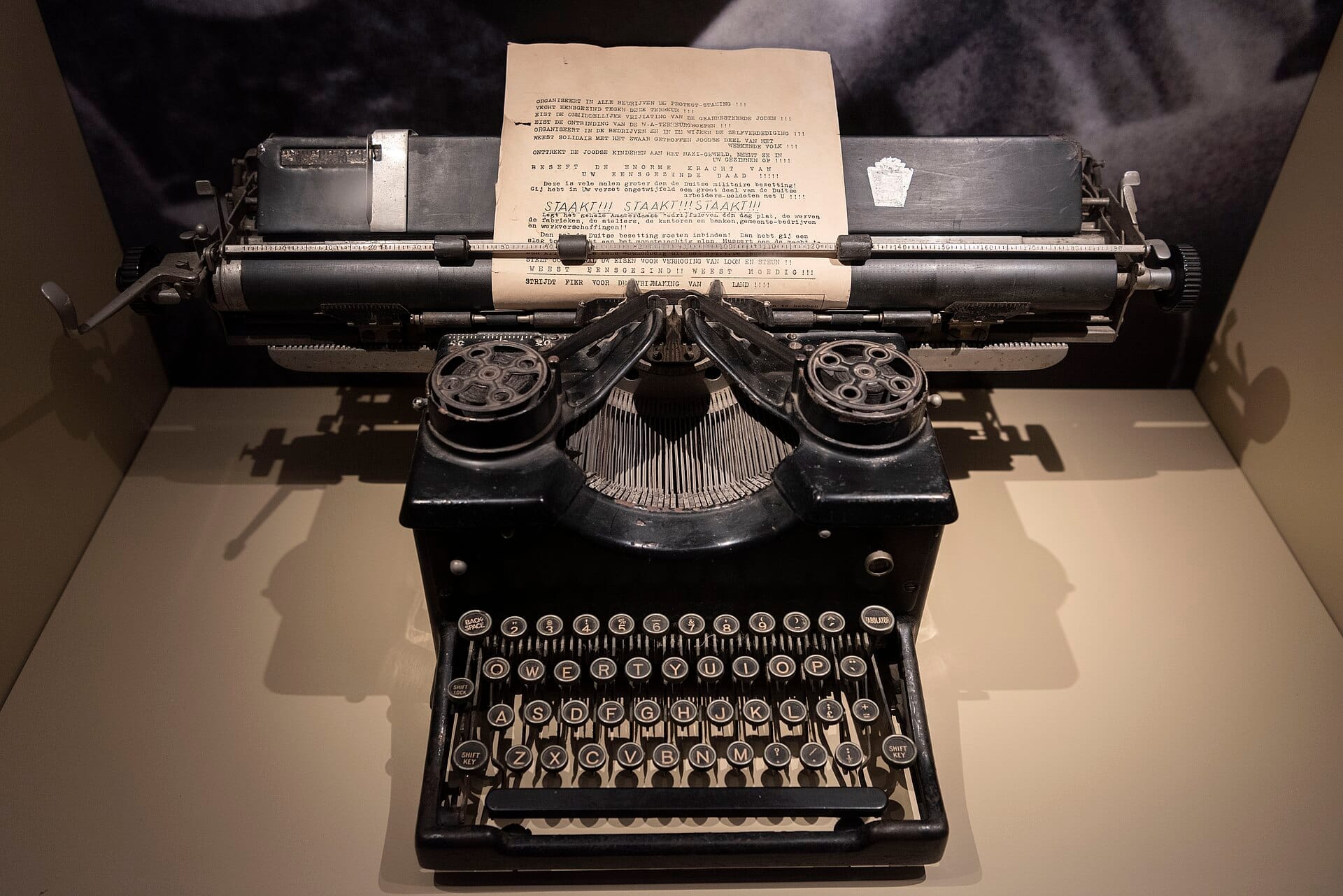 Máquina de escribir negra vintage con un documento de resistencia mecanografiado en exhibición en el Museo de la Resistencia Neerlandesa en Amsterdam, bien iluminada sobre un fondo neutro.