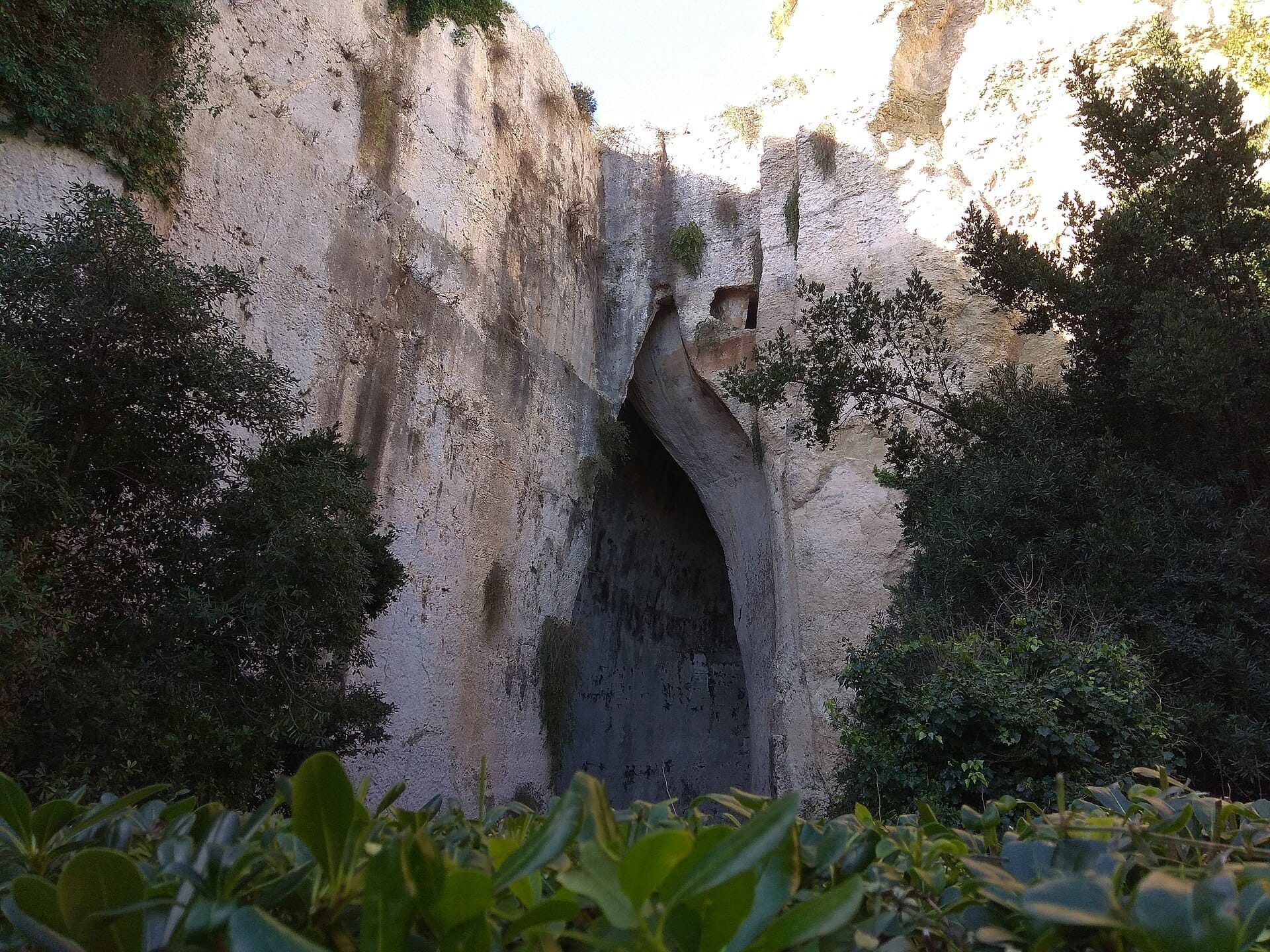 Eingang zum Ohr des Dionysius, einer hohen Kalksteinhöhle umgeben von üppigem Grün und verwitterten Felswänden in Syrakus, Sizilien.