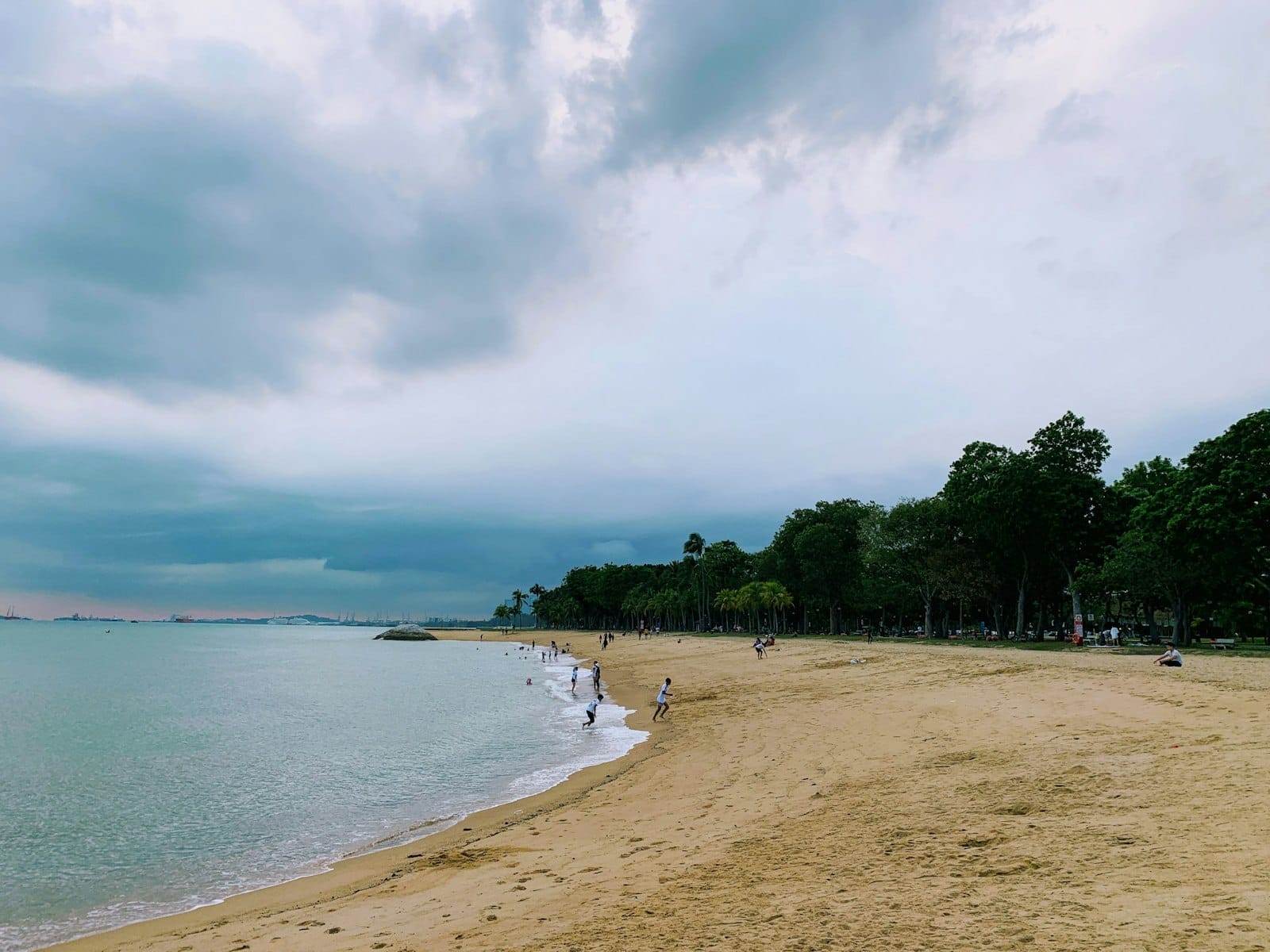 หาดทรายกว้างพร้อมคลื่นเบา ๆ ผู้คนเดินเล่นประปรายตามชายฝั่ง และต้นไม้เขียวชอุ่มใต้ท้องฟ้าเมฆครึ้มที่ East Coast Park สิงคโปร์