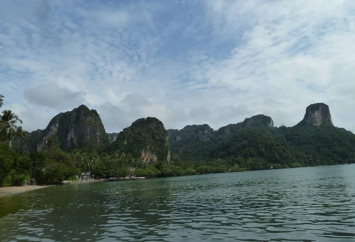 Panoramablick auf die Küstenlinie von East Railay Beach mit ruhigem, seichtem Wasser, Mangroven und Kalksteinfelsen im Hintergrund unter einem teils bewölkten Himmel.