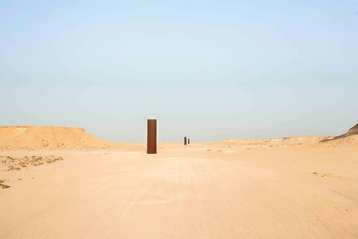 Weitwinkelperspektive auf Richard Serras Skulptur East-West/West-East in Katars Wüste – hohe Stahlplatten stehen in einer weiten Sandlandschaft unter klarem Himmel.