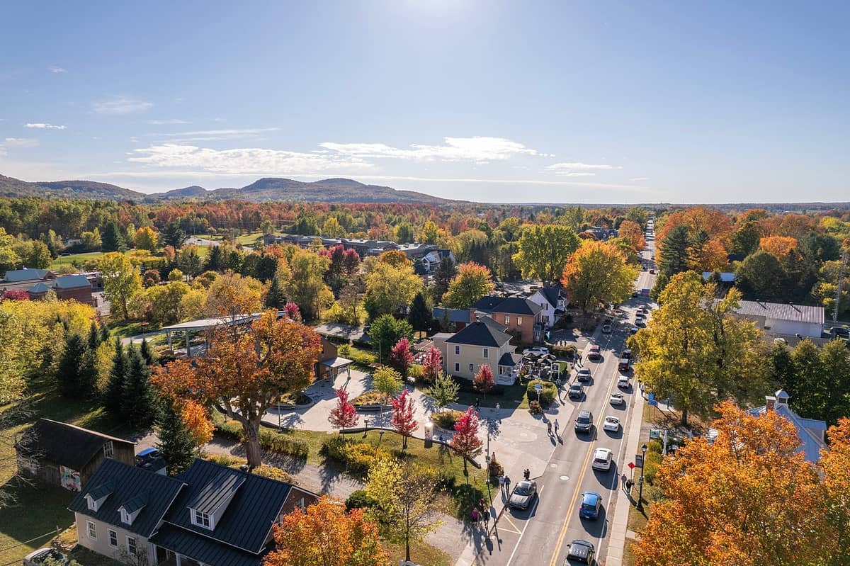 Luftaufnahme einer malerischen Stadt in den Eastern Townships mit Herbstbäumen, Häusern und einer Straße, die sich durch die bunte Landschaft erstreckt.