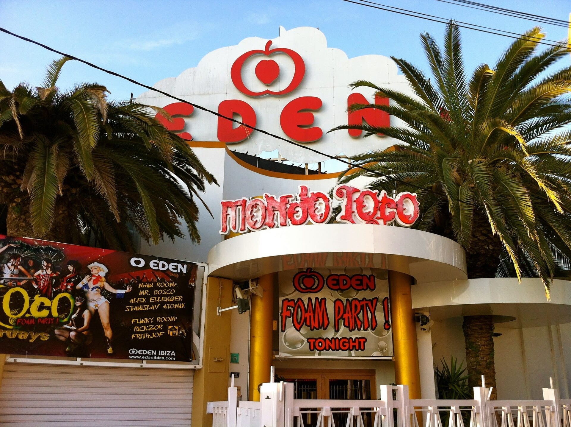 Ingresso del superclub Eden Ibiza con insegna rossa in grassetto, palme, striscione della festa della schiuma e architettura curva bianca sotto un cielo azzurro limpido.