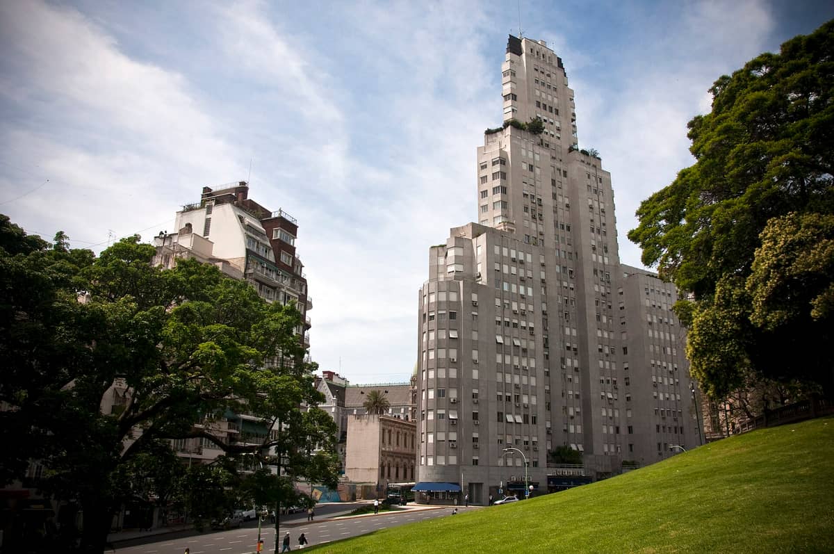 อาคารระฟ้า Edificio Kavanagh สไตล์ Art Deco สูงเด่นเหนือยอดไม้และตึกข้างๆ มองเห็นจาก Plaza San Martín ในวันแดดจ้ากับสนามหญ้าสีเขียวด้านหน้า