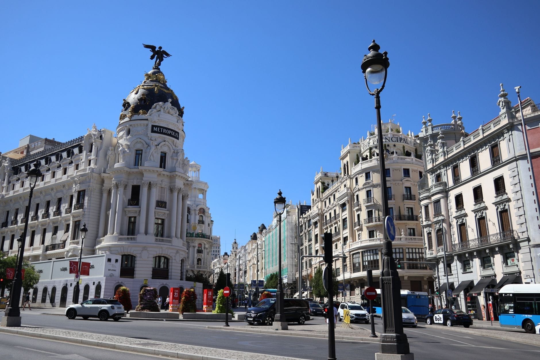 Vista grandangolare dell'Edificio Metrópolis all'angolo tra Calle de Alcalá e Gran Vía a Madrid sotto un cielo azzurro e limpido, con la cupola ornata e il vivace contesto urbano circostante.