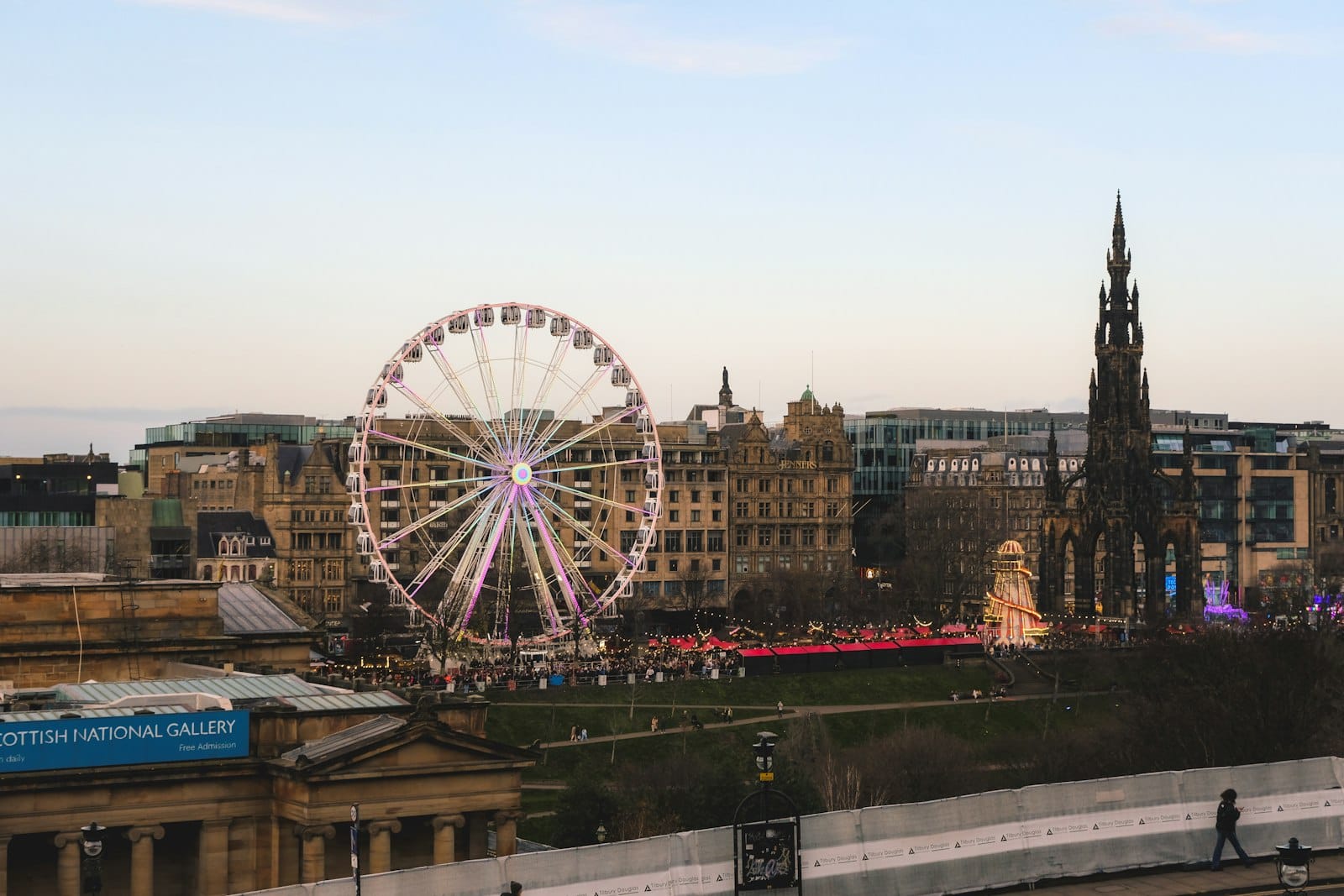 Vista degli East Princes Street Gardens di Edimburgo con il mercatino di Natale, la ruota panoramica, lo Scott Monument e gli edifici della città al tramonto.