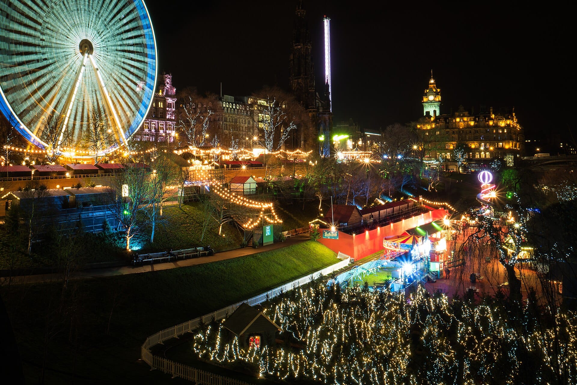 Ein weiter Nachtblick auf den Edinburgher Weihnachtsmarkt in den Princes Street Gardens mit einem hell erleuchteten Riesenrad, festlichen Marktständen, funkelnden Lichtern und historischen Gebäuden im Hintergrund.