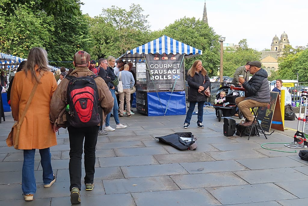 Besucher stöbern an Ständen unter blau-weiß gestreiften Zelten auf dem Edinburgh Farmers' Market, im Hintergrund Bäume und historische Gebäude.