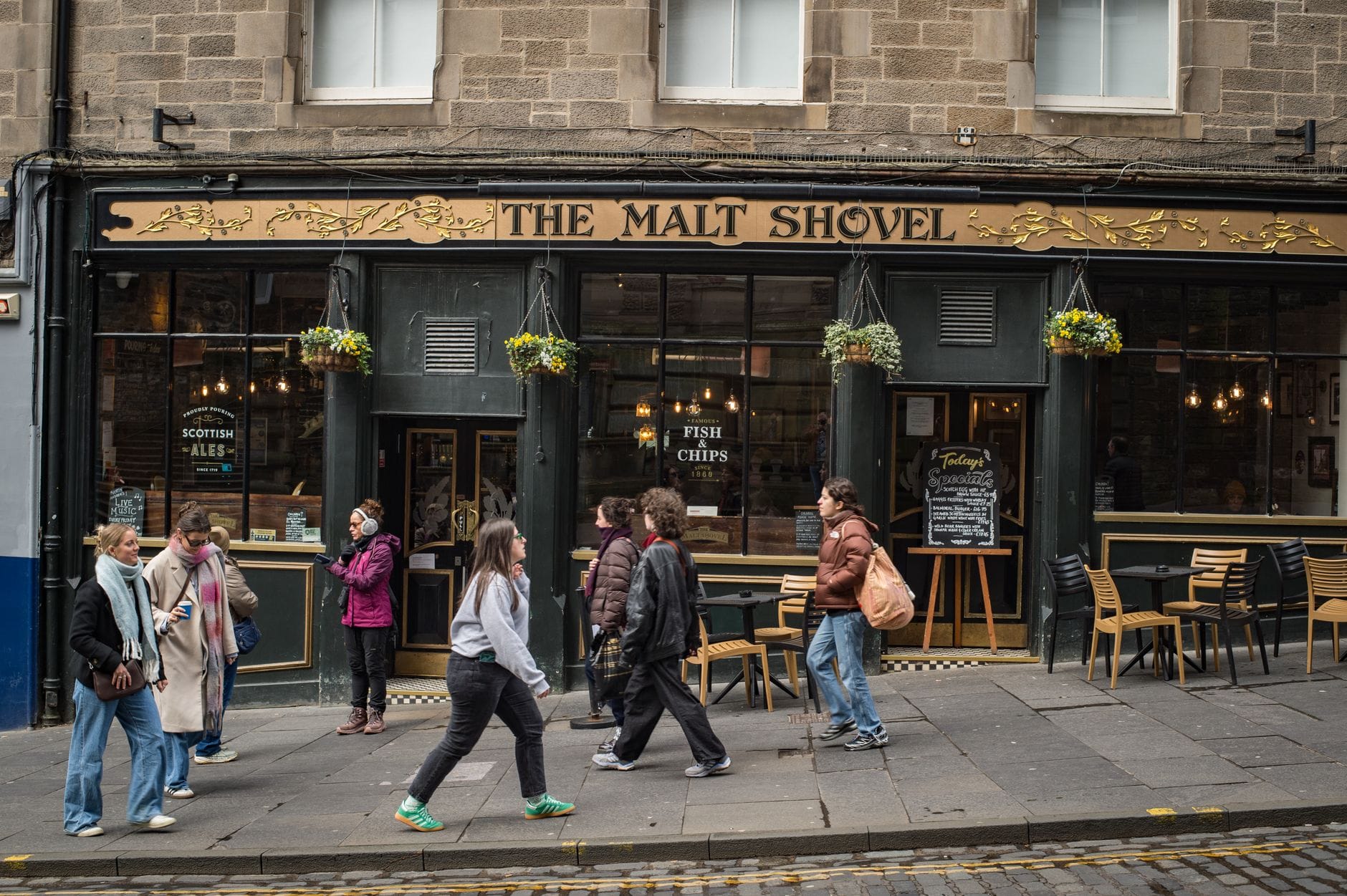 Straßenansicht des Pubs The Malt Shovel in Edinburgh mit vorbeigehenden Menschen, Pub-Schildern und Tischen draußen.