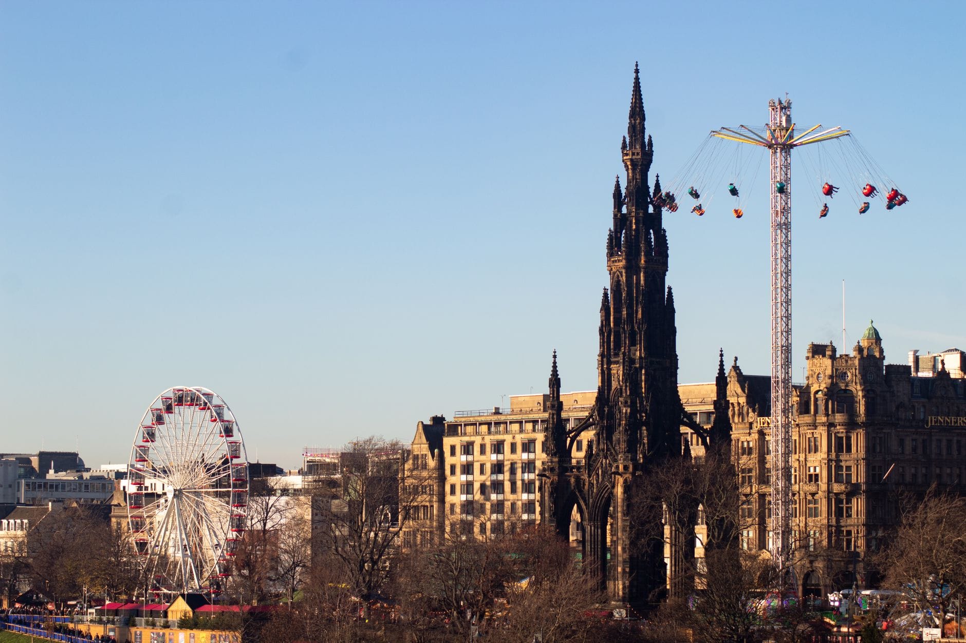 Panorama di Edimburgo con lo Scott Monument, una ruota panoramica e giostre allestite per le festività stagionali sotto un cielo azzurro limpido.