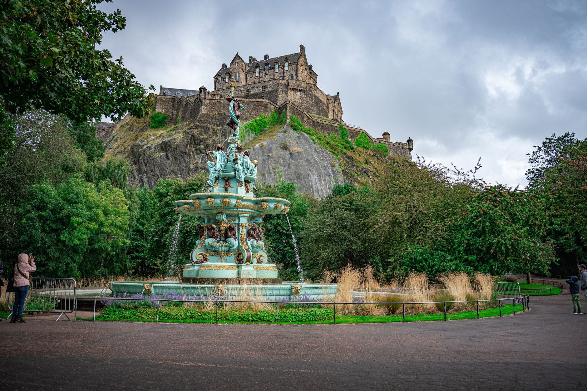 Vista ampia dell'Edinburgh Castle in cima a Castle Rock con l'ornata Ross Fountain in primo piano, circondata da alberi e vialetti.