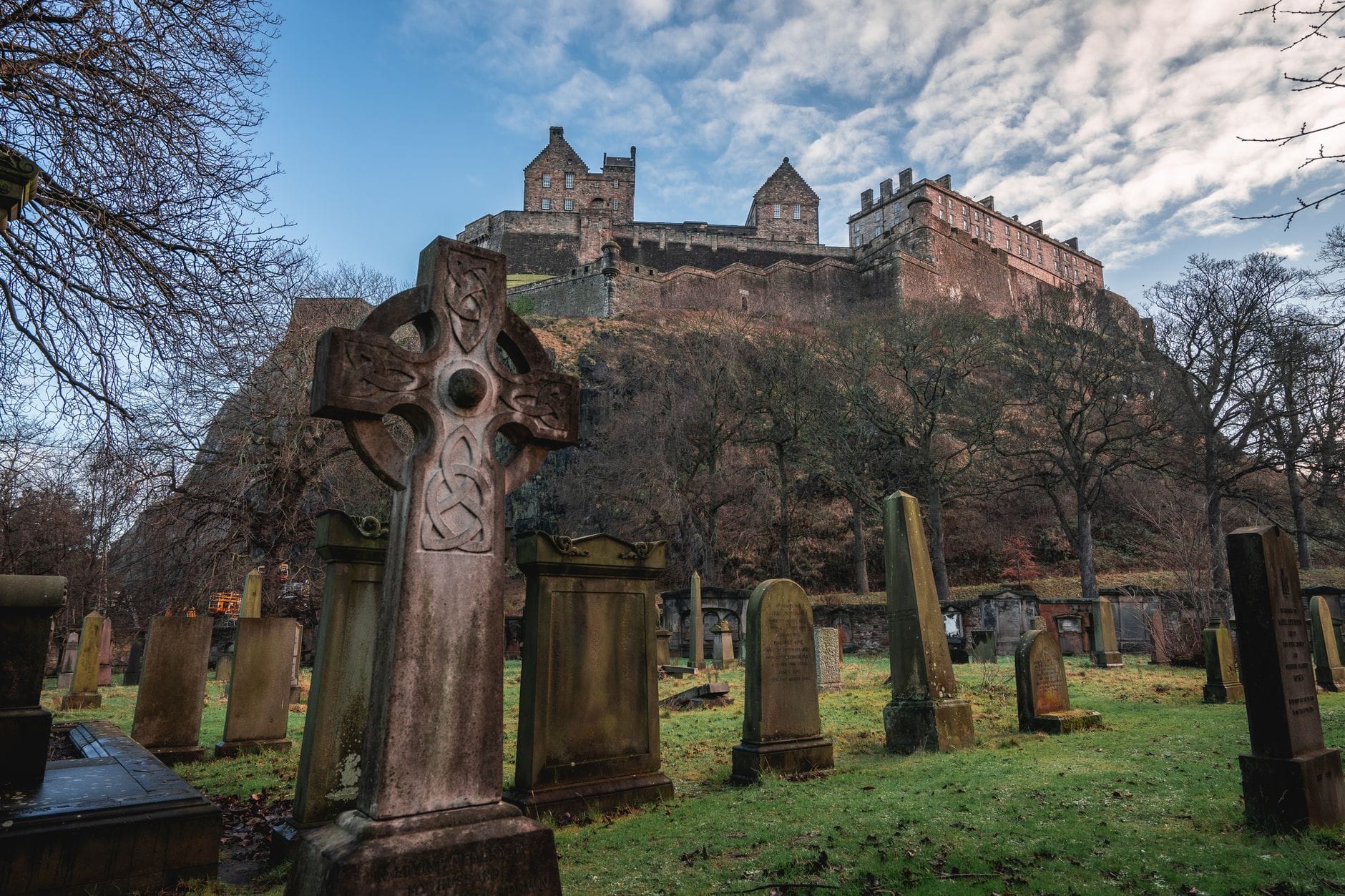 Blick auf historische Grabsteine eines Kirchhofs unterhalb des Edinburgh Castle, mit kahlen Bäumen und einem bedrohlichen Himmel, der eine unheimliche Atmosphäre erzeugt.