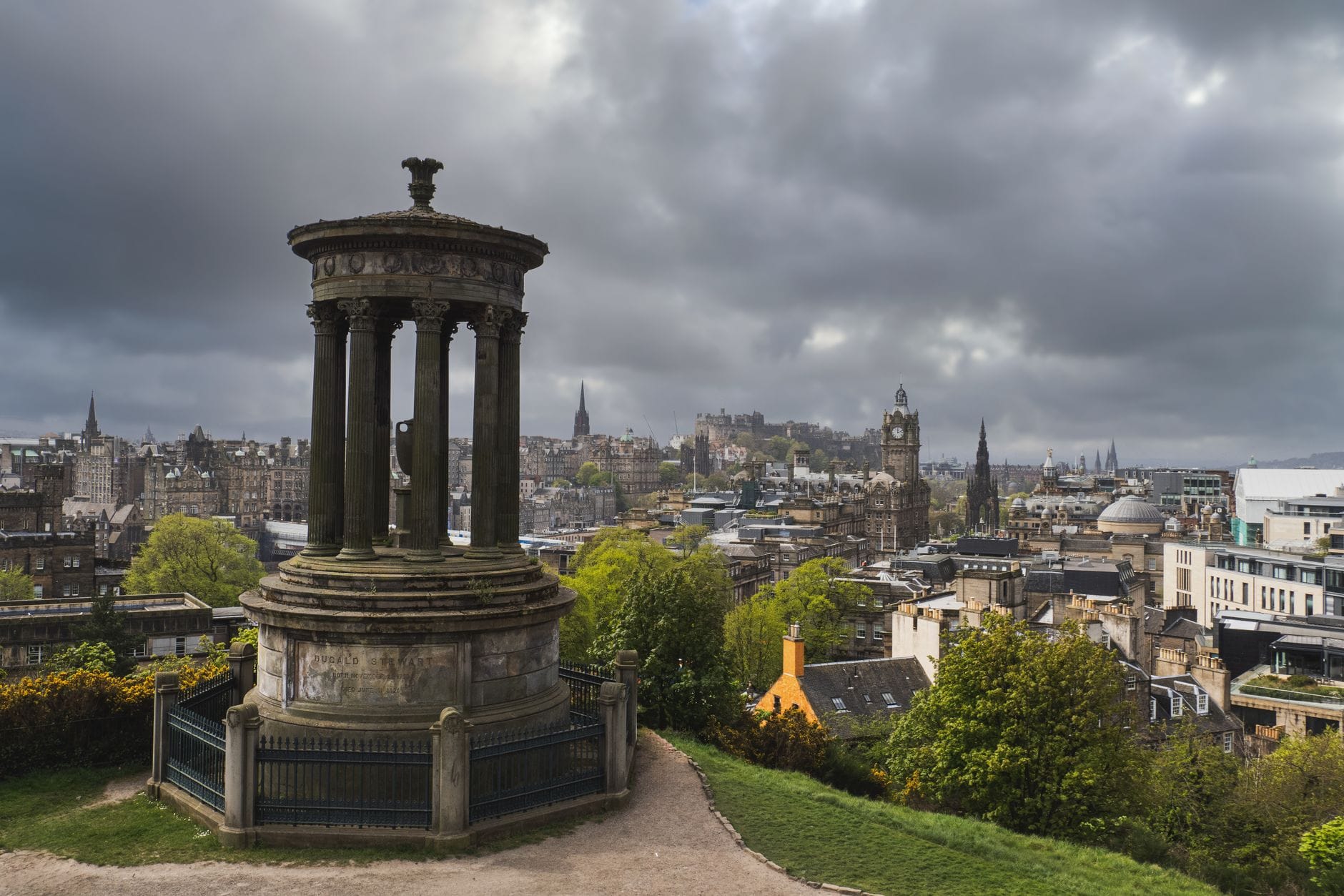 Düsterer Blick vom Calton Hill über die Skyline von Edinburgh mit dem Dugald Stewart Monument und dunklen Wolken darüber, der die gotische und atmosphärische Landschaft der Stadt hervorruft.