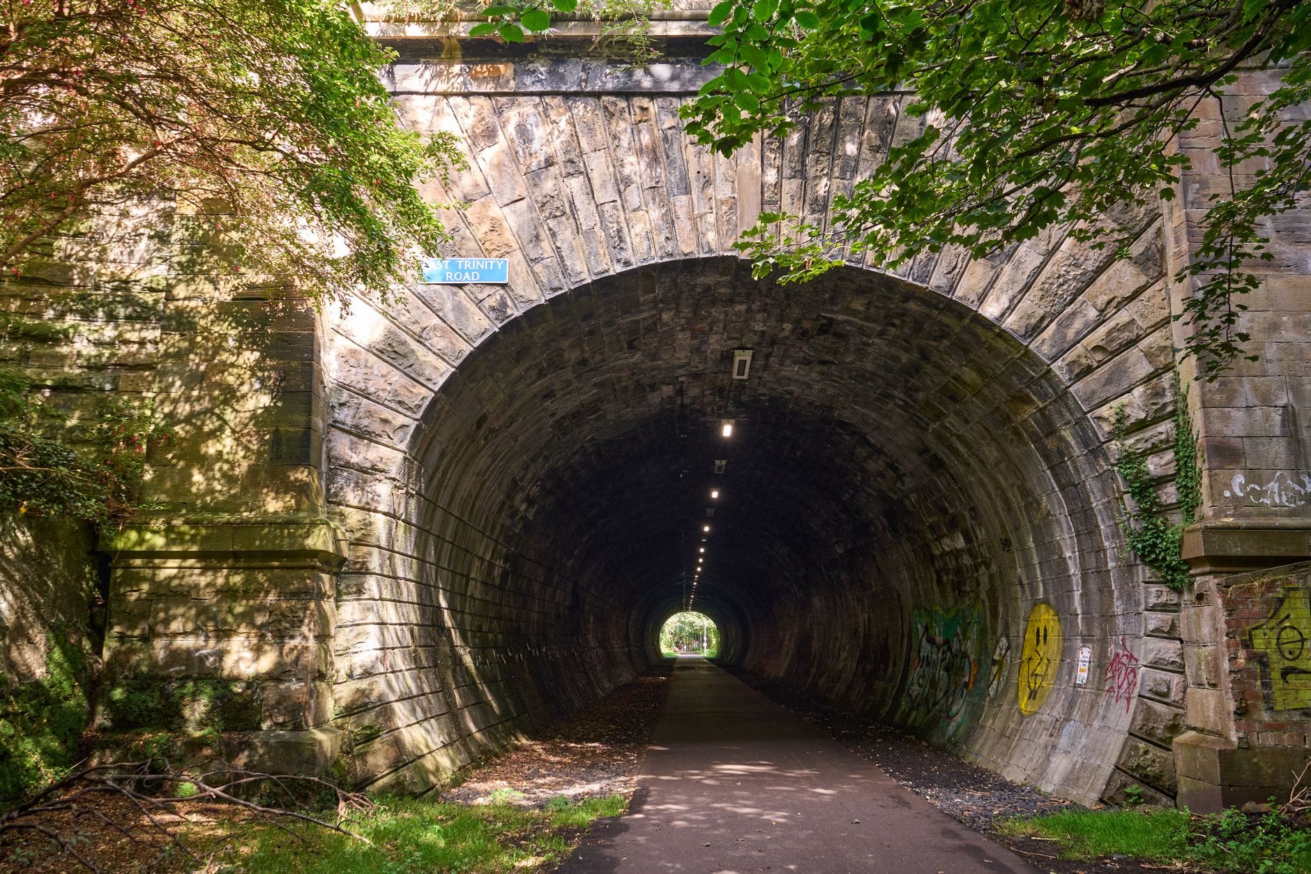 Ein steinerner Tunneleingang mit der Aufschrift Colinton Tunnel, umgeben von Grün, der in die Dunkelheit führt, mit einem beleuchteten Weg im Inneren.