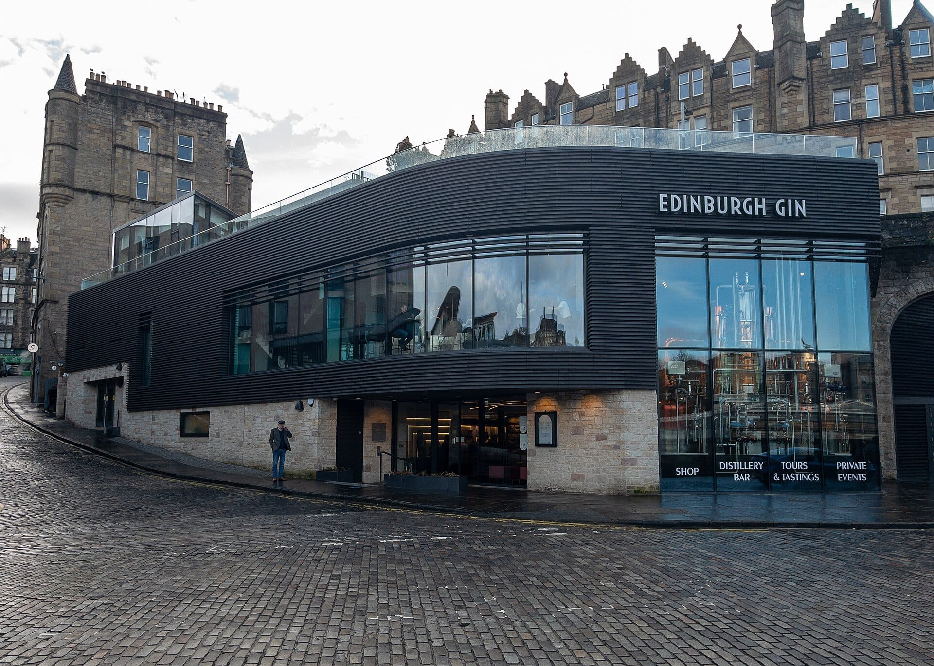 Modernes Äußeres der Edinburgh Gin Distillery in The Arches, mit großen Glasfenstern und historischen Steingebäuden im Hintergrund auf einer Kopfsteinpflasterstraße.