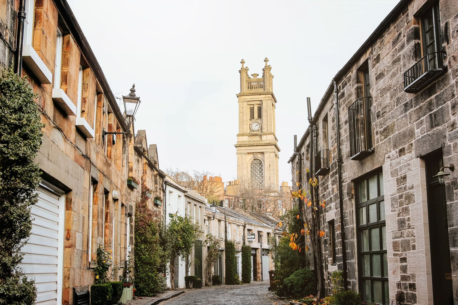 Kopfsteingepflasterte Mews-Straße in Edinburgh, gesäumt von malerischen Steinhäuschen und Grünpflanzen, mit dem Uhrenturm der St. Stephen's Church über den Dächern.