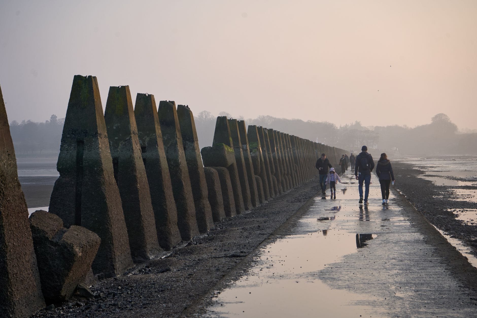 Menschen, die bei Ebbe entlang der beeindruckenden Betonpfeiler des Damms von Cramond Island spazieren, mit nassem Sand und einem nebligen Hintergrund.