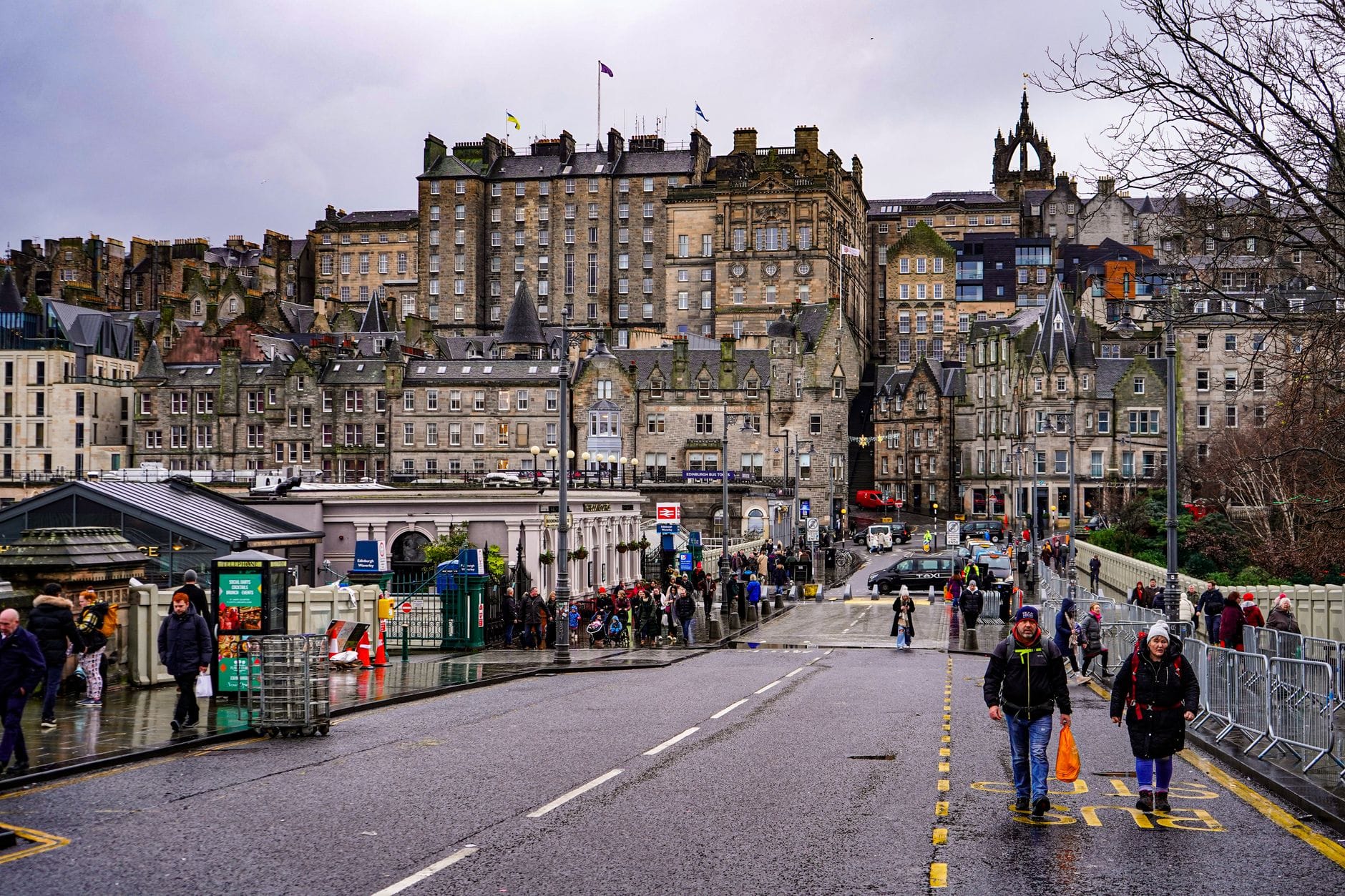 Blick auf das Zentrum von Edinburgh mit nassen Straßen, Menschen in Mänteln und historischen Steingebäuden unter einem grauen Himmel.