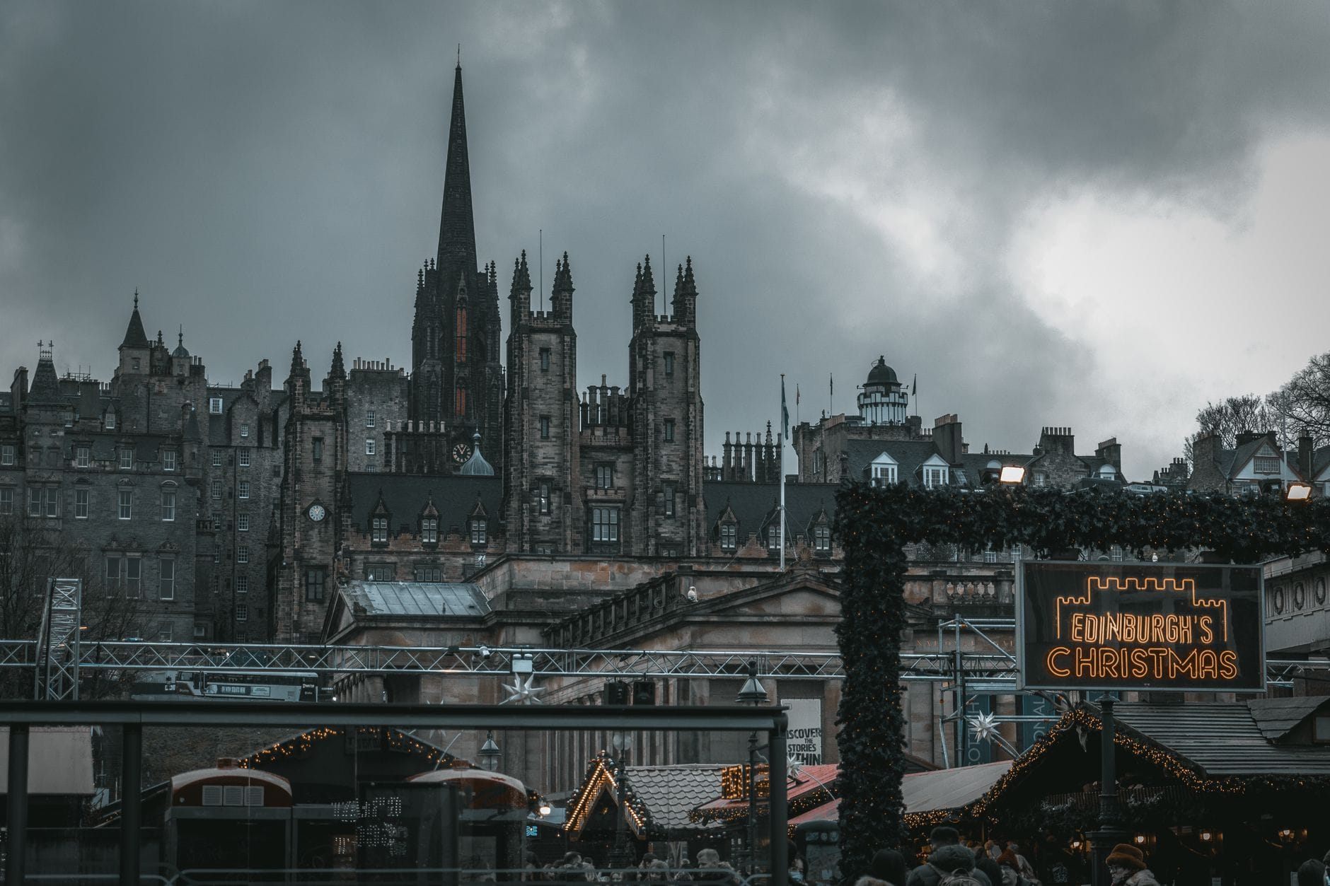 Blick auf den Weihnachtsmarkt von Edinburgh mit festlichen Ständen, Menschenmassen, gotischer Architektur und einem beleuchteten Schild 'Edinburgh's Christmas' unter bewölktem Himmel.