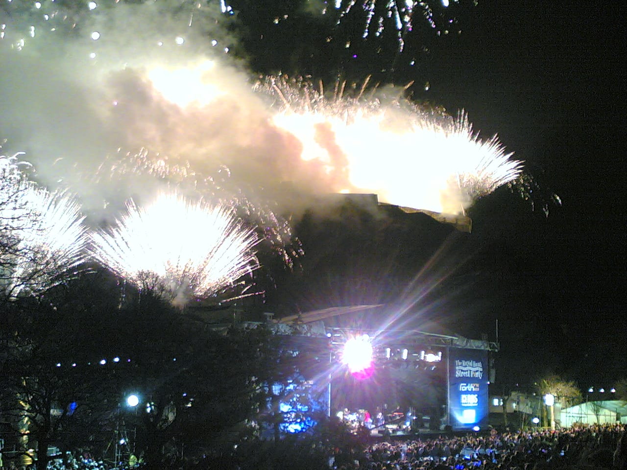 Große Menschenmenge bei Nacht mit Feuerwerk über Edinburgh Castle und einer beleuchteten Bühne während der Edinburgh Hogmanay Straßenfestfeierlichkeiten.
