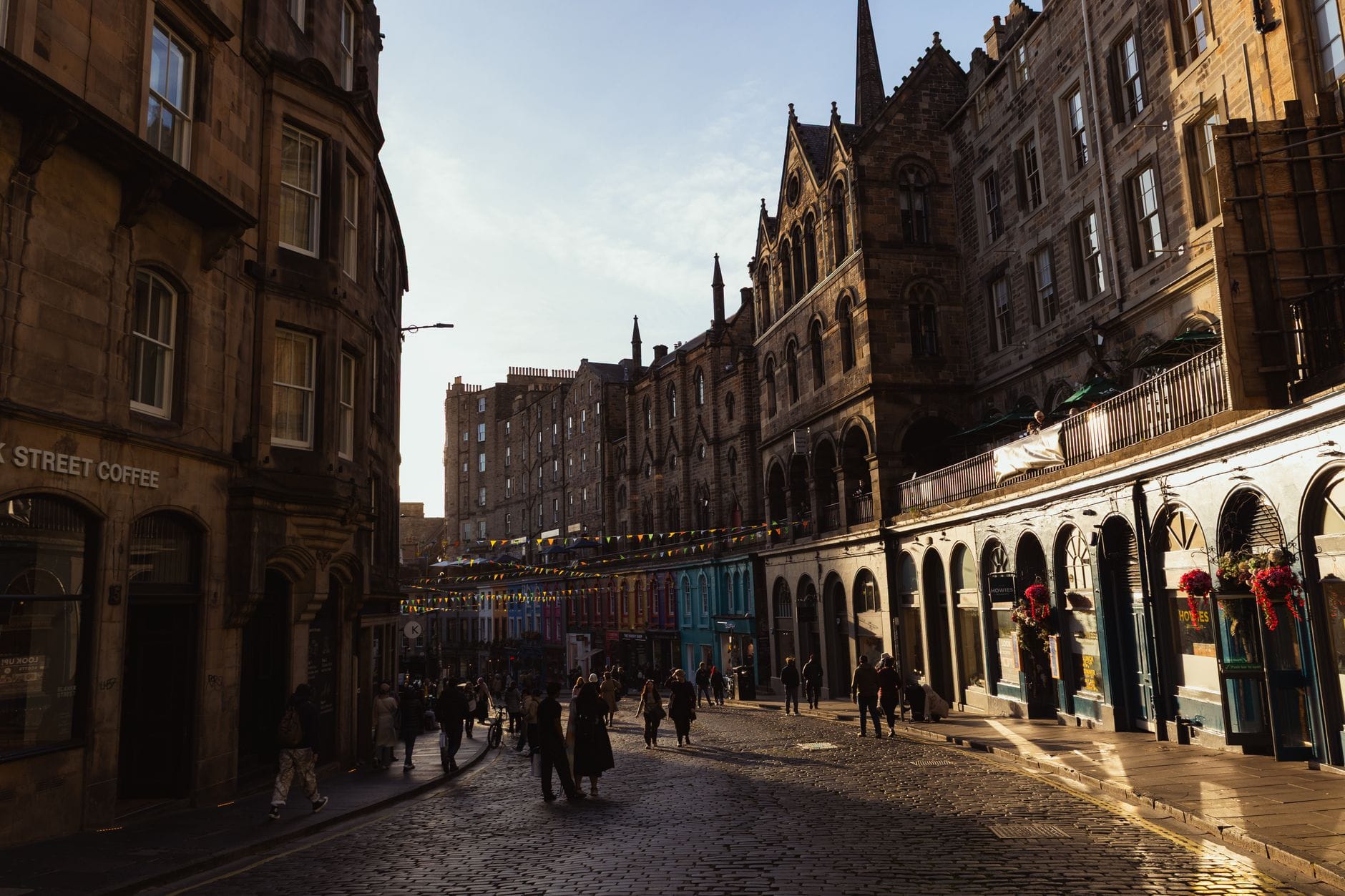 วิวตามถนน Victoria Street ในเมืองเอดินบะระมุ่งสู่ Cowgate พร้อมอาคารหินประวัติศาสตร์ ร้านค้าสีสันสดใส ธงประดับเหนือถนน และผู้คนที่เดินอยู่บนถนนคึกคัก