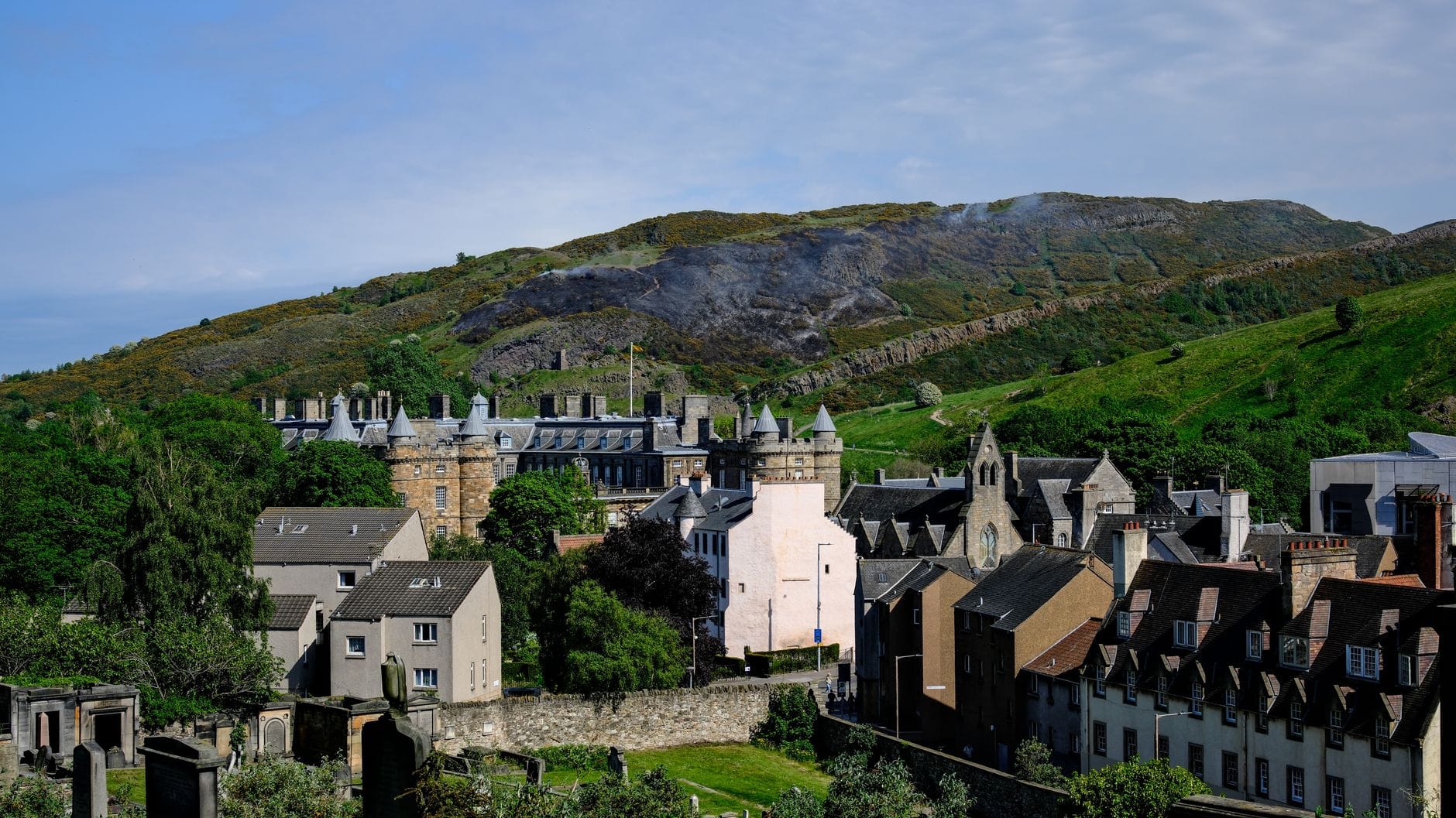 Vista panoramica del Palace of Holyroodhouse con i circostanti edifici storici e Arthur's Seat che si innalza sullo sfondo, sotto un cielo azzurro.