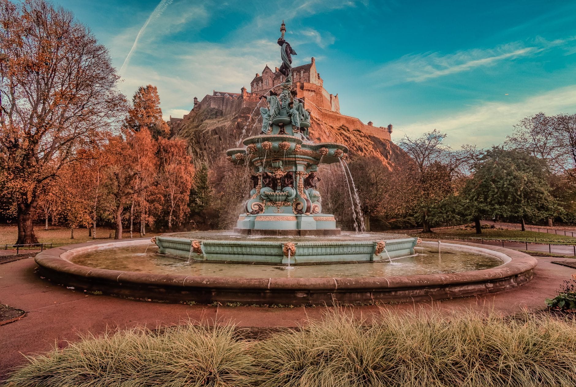 Vista della Ross Fountain nei Princes Street Gardens con l'Edinburgh Castle sulla collina sullo sfondo, alberi frondosi e cielo azzurro.