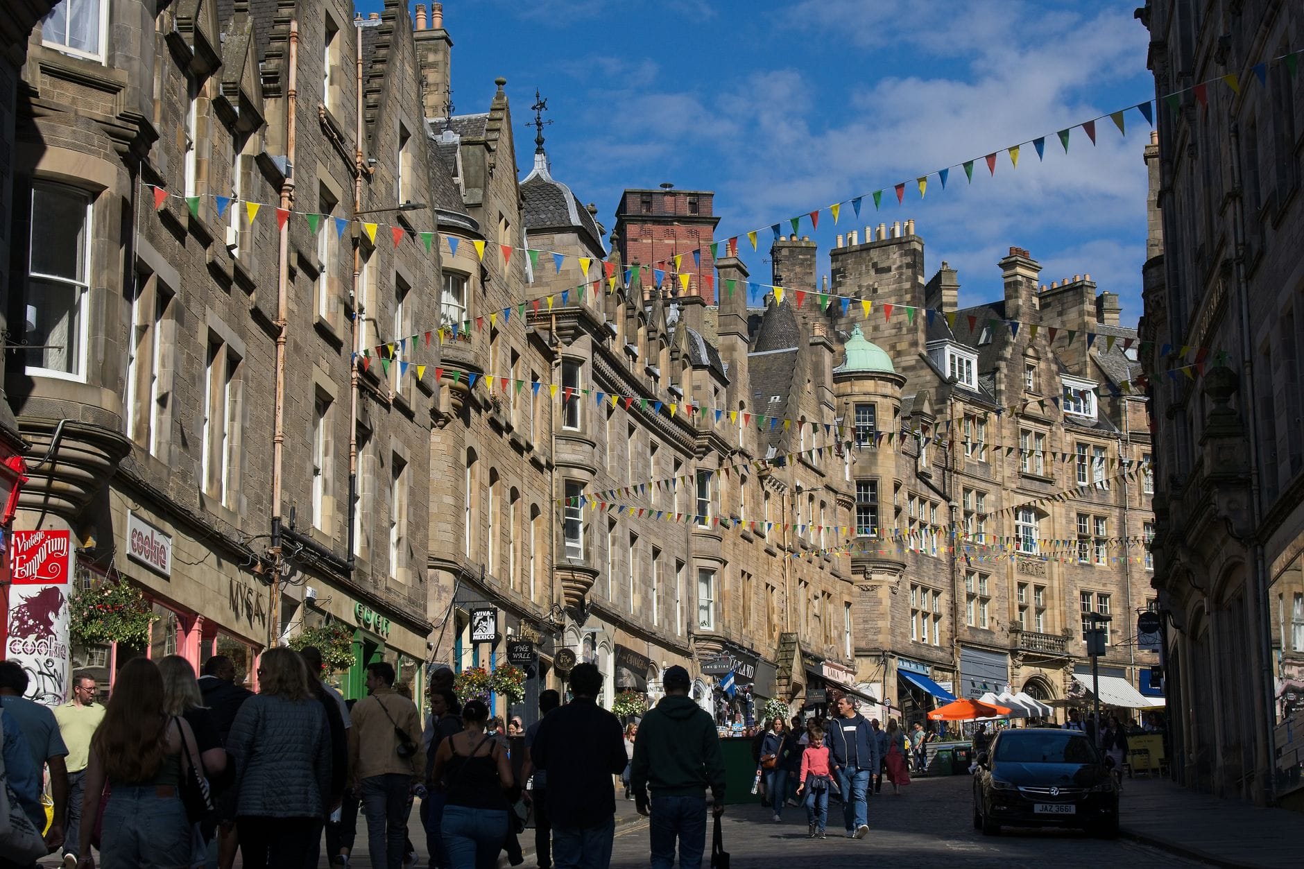 Eine belebte Straße in Edinburgh mit historischen Steingebäuden und bunten Wimpelketten unter blauem Himmel, Menschen schlendern und genießen die lebhafte Sommeratmosphäre.
