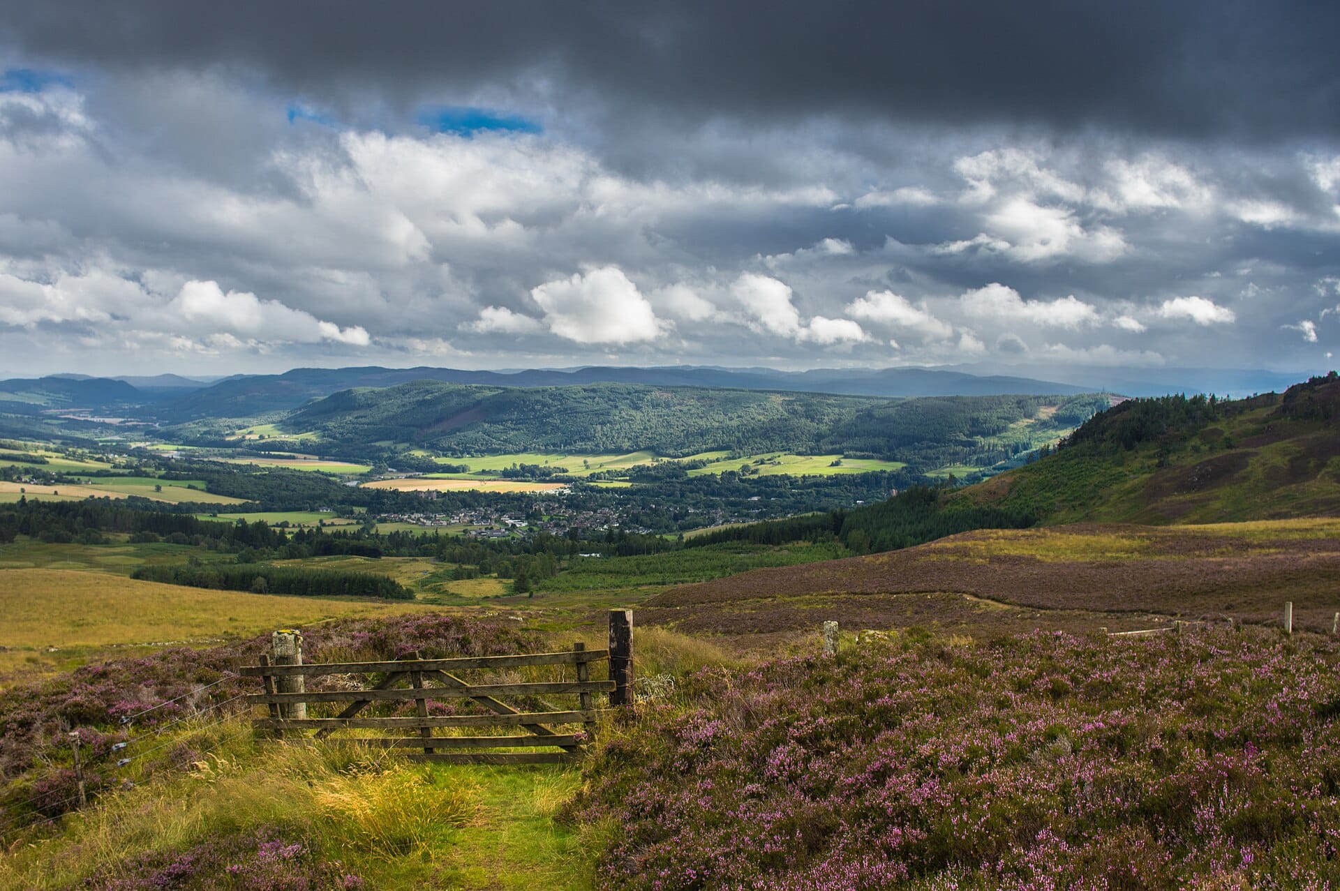 Weitläufige Landschaftsansicht über die Highlands mit einem Tor und Heidekraut im Vordergrund sowie einer Talstadt, umgeben von Hügeln und dramatischen Wolken.