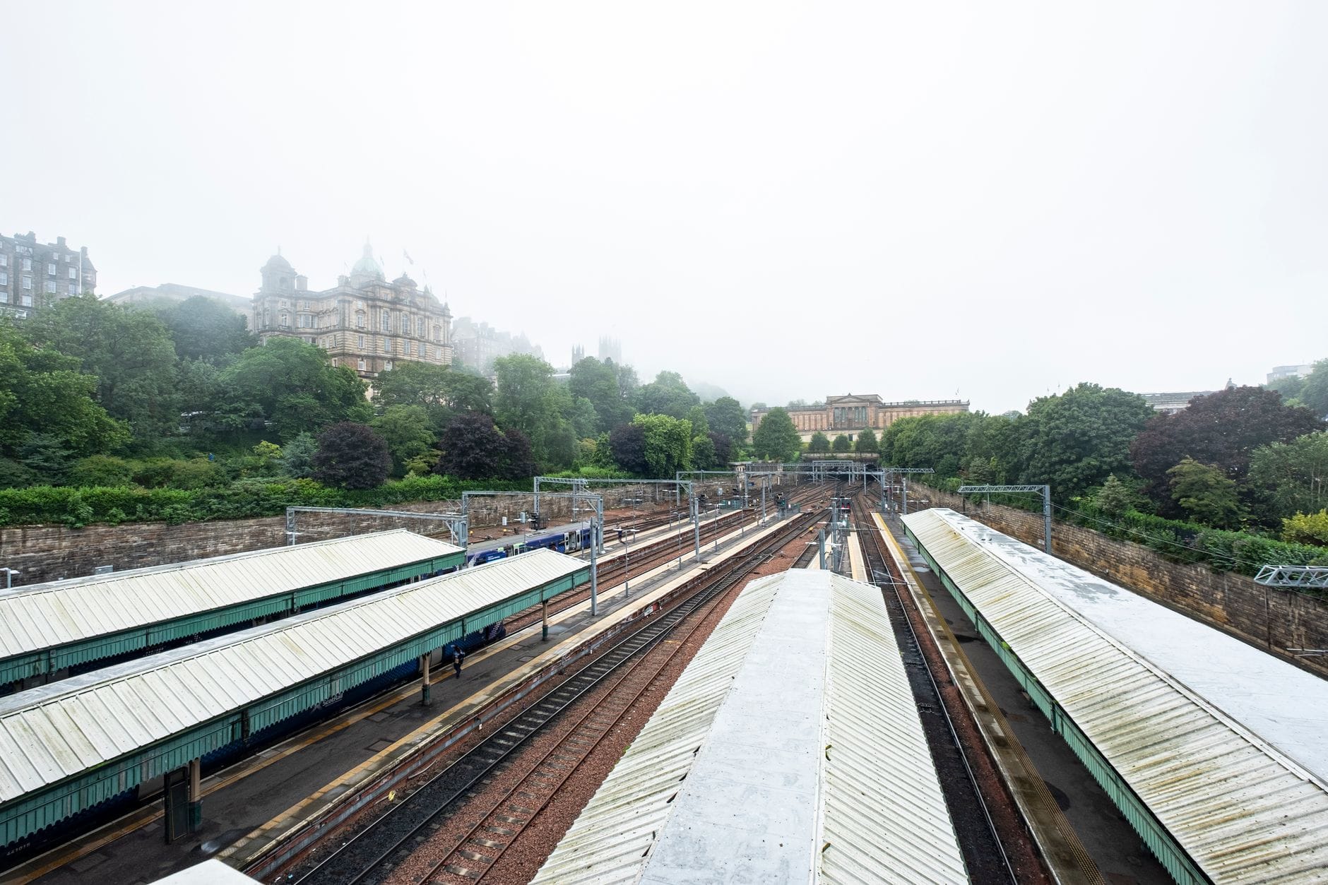 Weitwinkelansicht der Bahnsteige des Bahnhofs Edinburgh Waverley, mit Gleisen, die sich in die Ferne erstrecken, und historischen Gebäuden im Hintergrund.