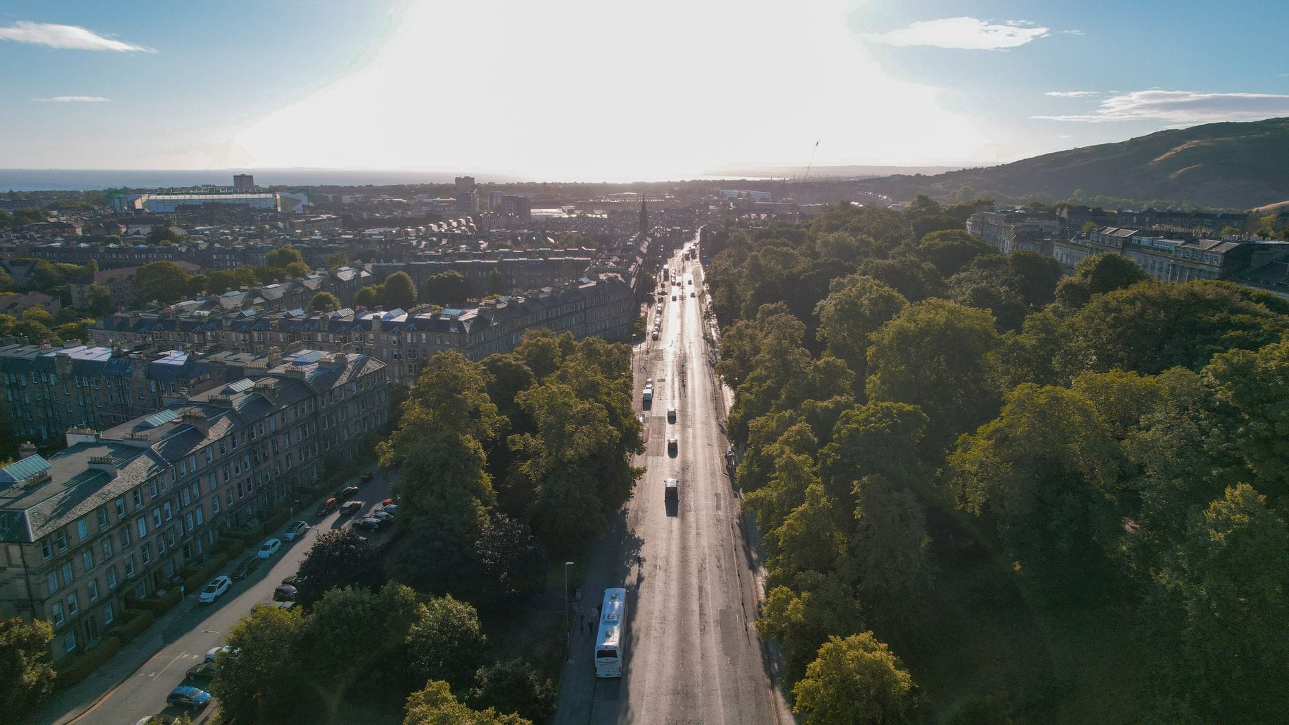 Eine breite, sonnenbeschienene Straße, die aus der Stadt Edinburgh herausführt, gesäumt von klassischen Gebäuden und laubbedeckten Bäumen, mit Hügeln im Hintergrund und blauem Himmel darüber.