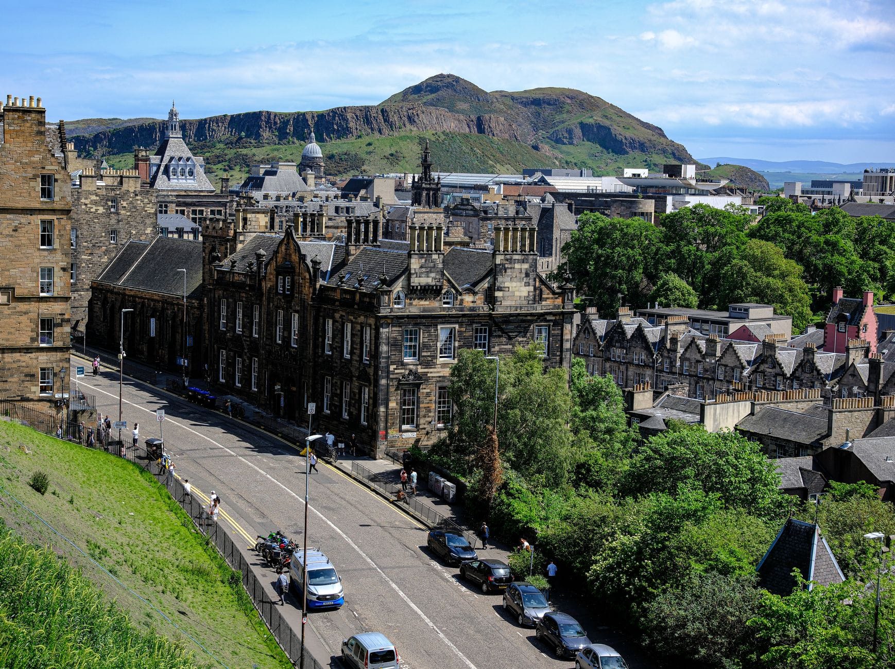 Ein klarer Blick über das Zentrum von Edinburgh bei Tag: historische Gebäude, Arthur's Seat im Hintergrund und eine begrünte Straße mit parkenden Autos im Vordergrund.