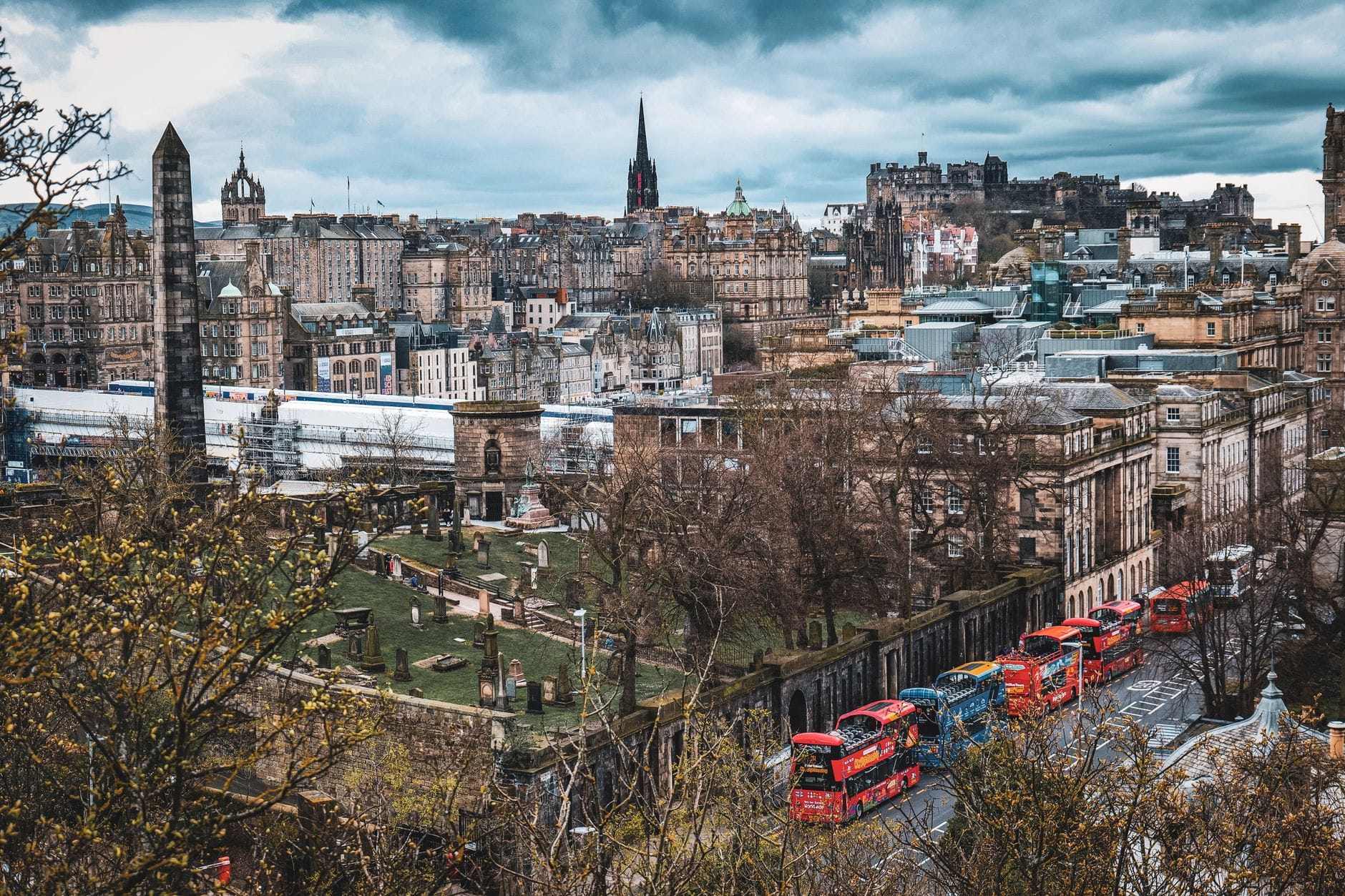Panoramablick auf Edinburghs historische Old Town mit markanten Steingebäuden, Kirchturmspitzen, Edinburgh Castle in der Ferne und roten Touristenbussen entlang einer Hauptstraße.