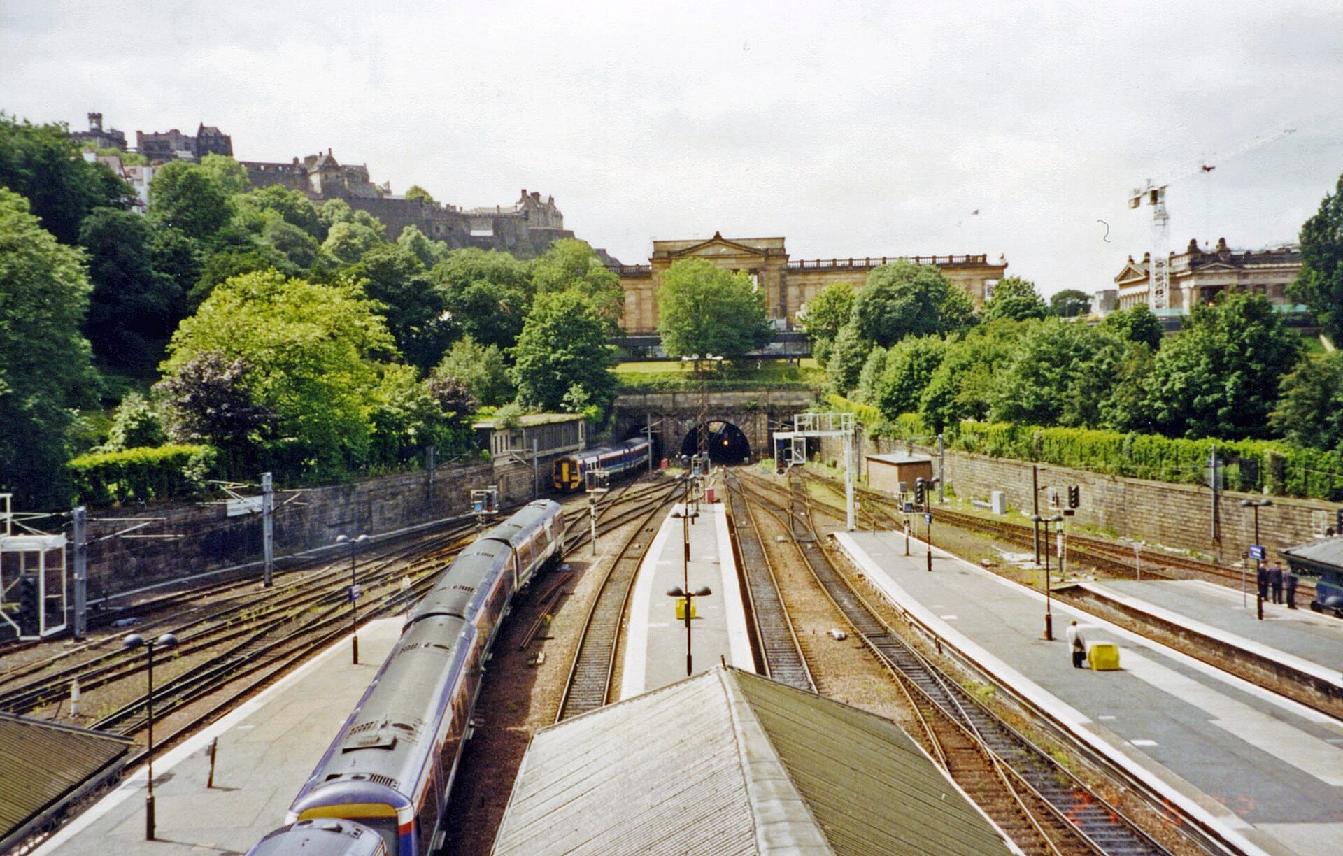 Gleise und Bahnsteige am Bahnhof Edinburgh Waverley, umgeben von Grünflächen, mit Gebäuden der Edinburgher Innenstadt im Hintergrund.