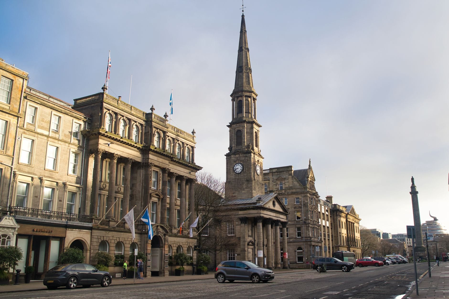 Klassische georgianische Architektur und ein Kirchturm im New Town von Edinburgh entlang einer breiten, eleganten Straße mit Steingebäuden und sichtbaren Flaggen.