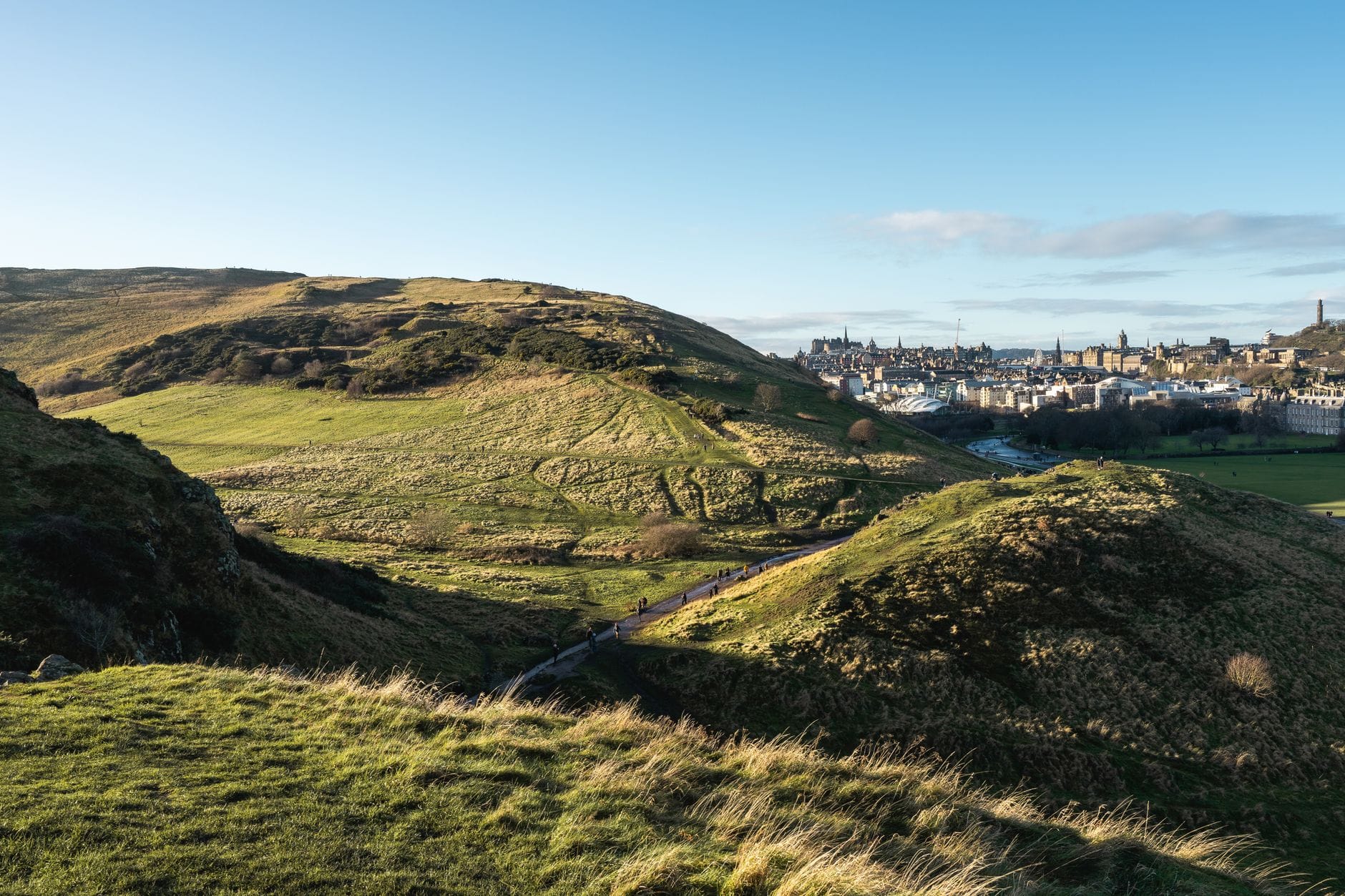 Sanfte grüne Hügel und ein Fußweg im Holyrood Park mit der Skyline von Edinburgh und Sehenswürdigkeiten im Hintergrund unter blauem Himmel.